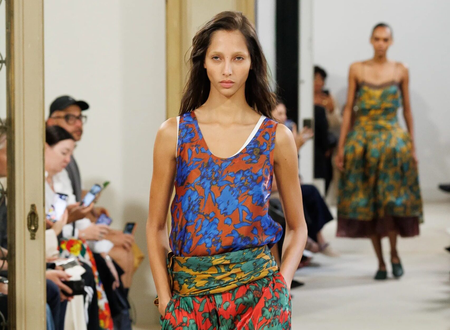 Modelo desfilando con una blusa sin mangas estampada en tonos azules y naranjas, fajín multicolor en la cintura y pantalones bombachos rojos con motivos verdes.
