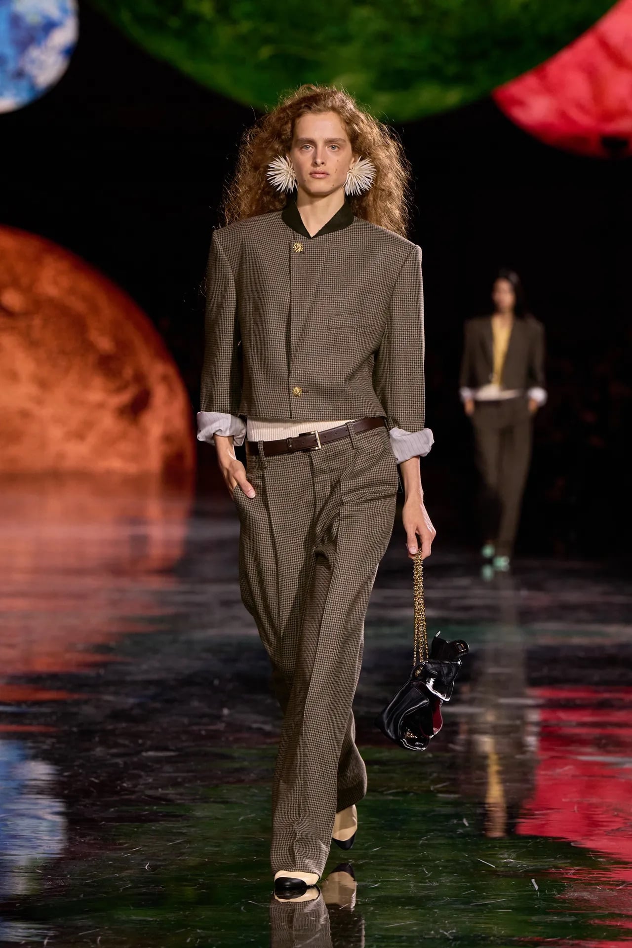 Modelo desfilando con un conjunto sastre de chaqueta corta estructurada y pantalones anchos de cuadros sutiles, luciendo aretes llamativos y un bolso de mano negro con cadena dorada.