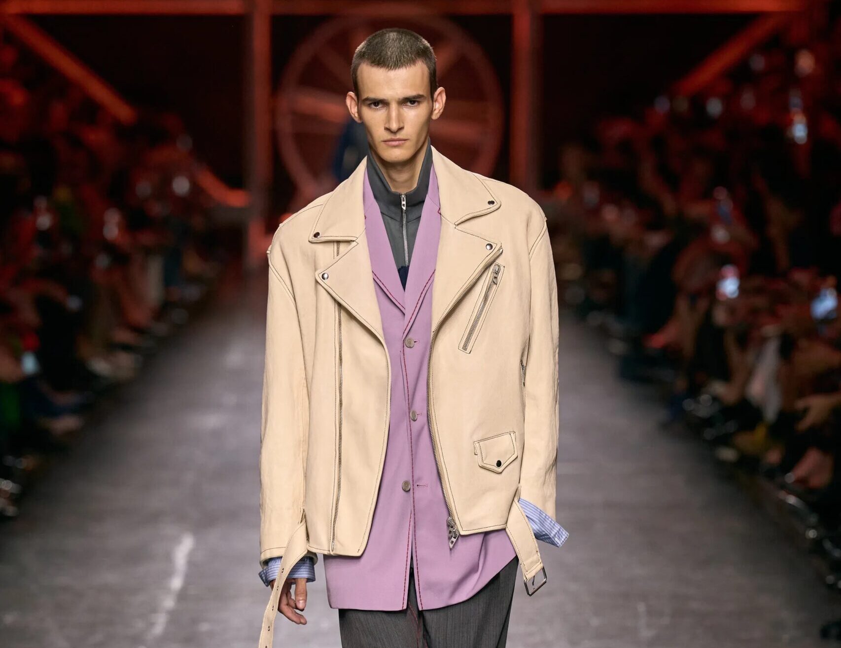 Modelo masculino con chaqueta motera de piel en tono beige claro, sobre una camisa lila, saco formal y pantalón gris de traje.