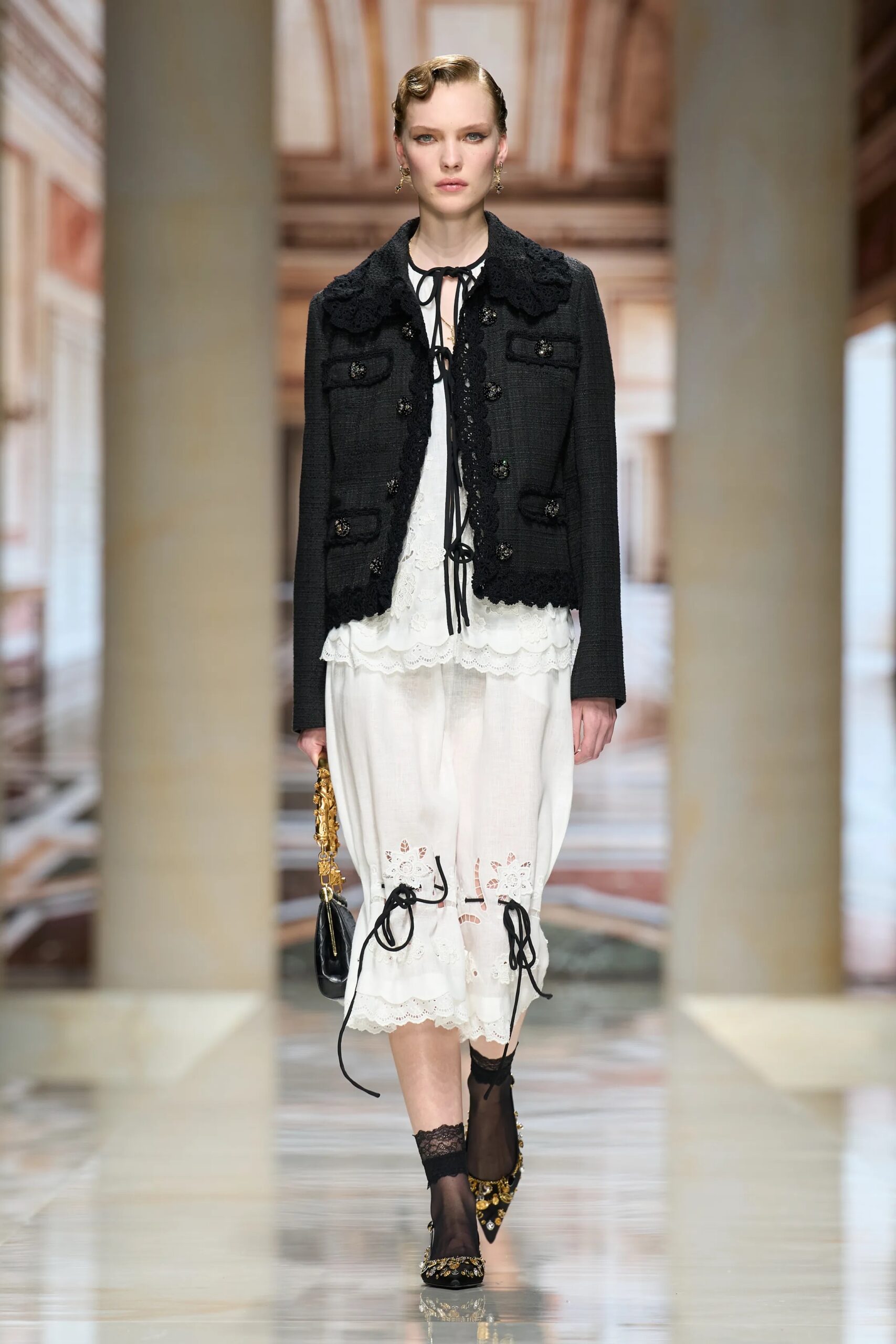 Modelo desfilando con una chaqueta corta de tweed negro con detalles texturizados y botones decorativos, sobre una blusa blanca de encaje con lazos y una falda midi vaporosa con detalles bordados, capturada por Isidore Mantag.