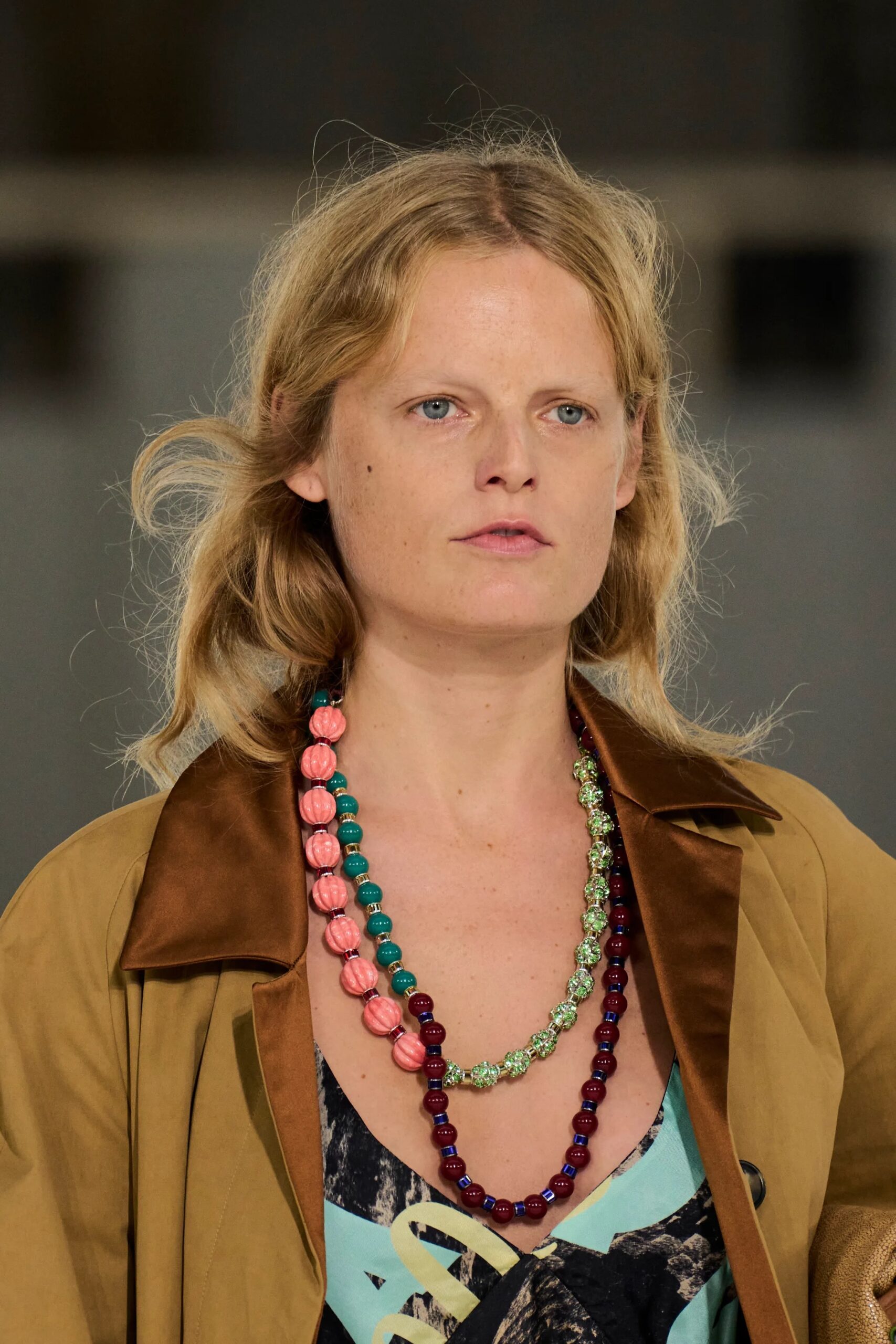 Primer plano de una modelo con collares superpuestos de cuentas coloridas en tonos rosa, verde y azul, complementados con una chaqueta de solapas marrones y un vestido estampado