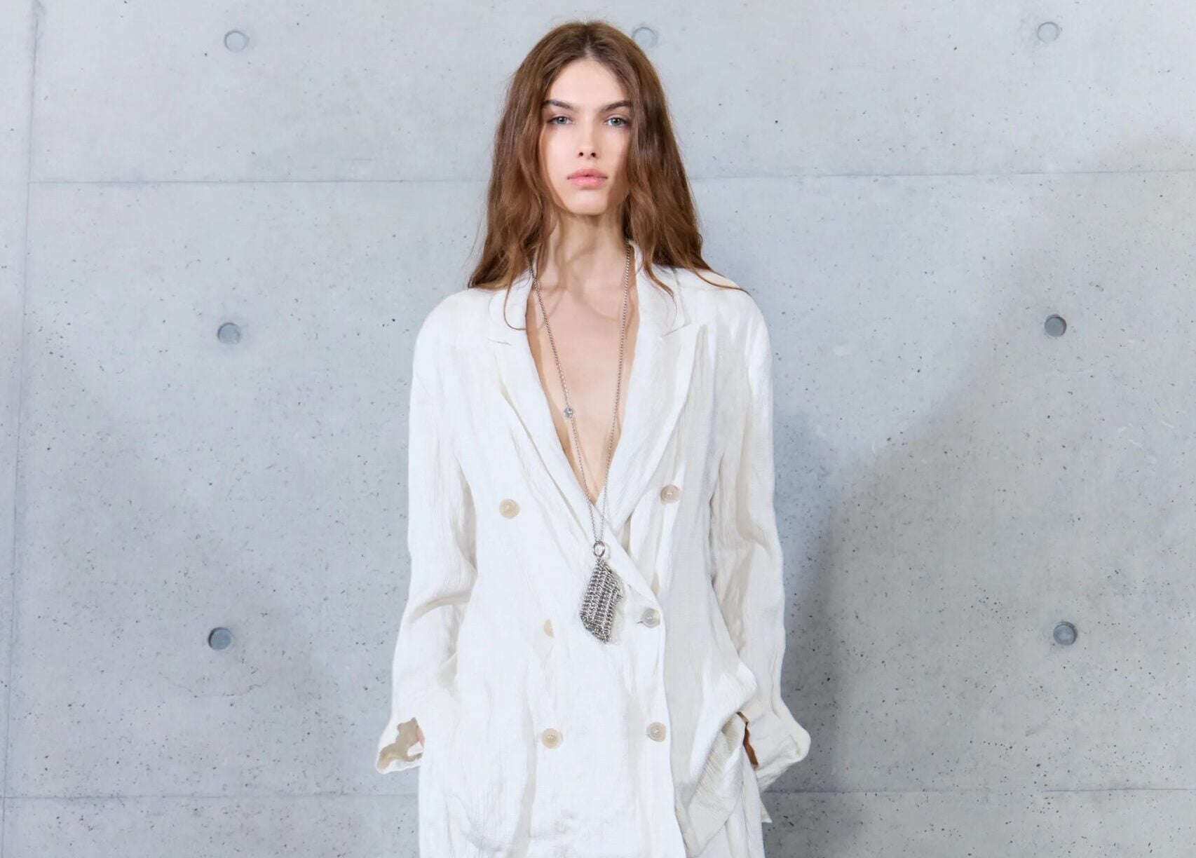 Modelo posando con un traje sastre fluido de doble botonadura en tono blanco roto, acompañado de collares largos metálicos y sandalias claras.