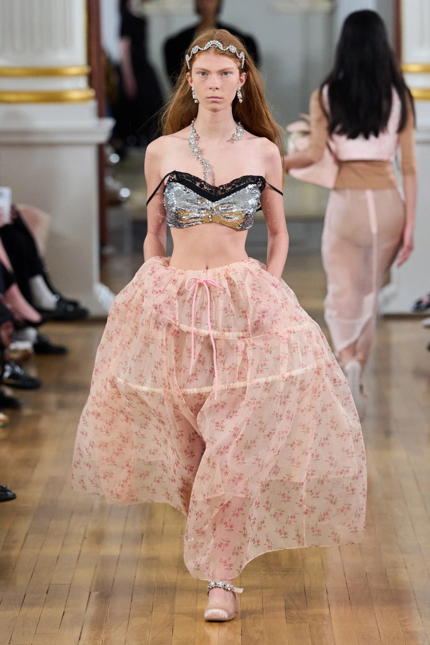 Modelo en pasarela con un top estilo bralette de lentejuelas plateadas y encaje negro, combinado con una falda midi transparente con estampado floral rosa y cordón ajustable.