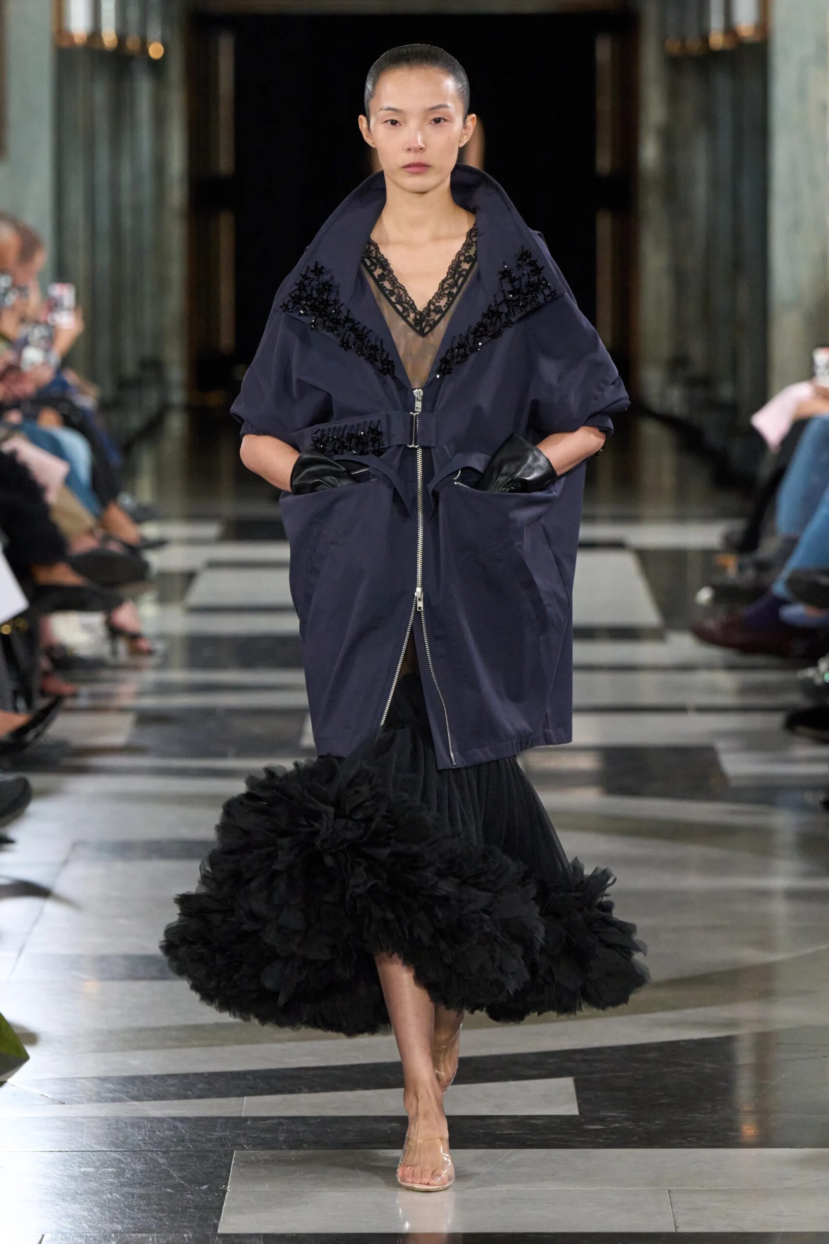 Modelo con un abrigo estructurado azul marino con detalles de encaje negro en el escote, falda abullonada de tul negro y guantes de piel.