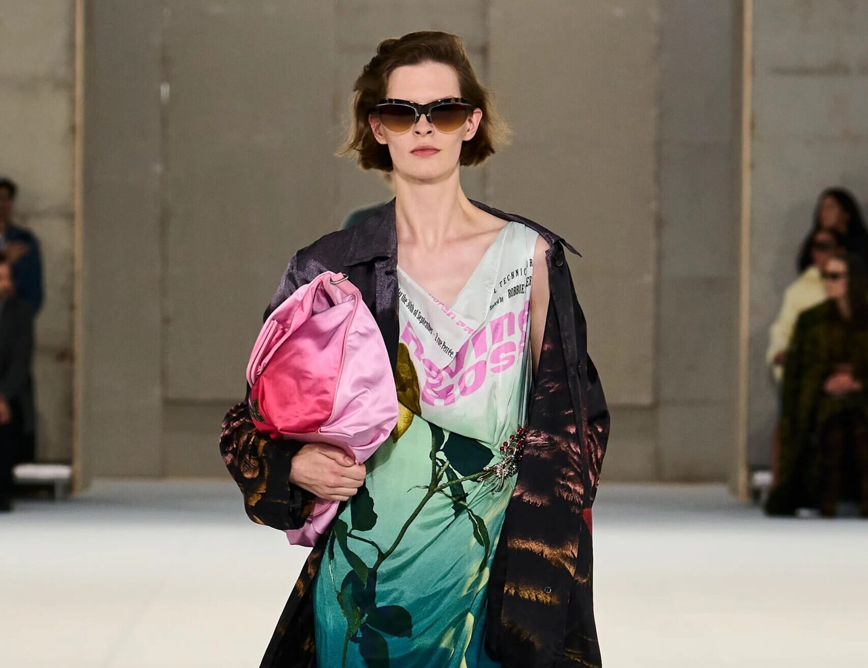 Modelo desfilando con un vestido midi estampado en tonos verdes y azules, cubierto por una gabardina larga oscura, gafas de sol y cargando un bolso rosa brillante.