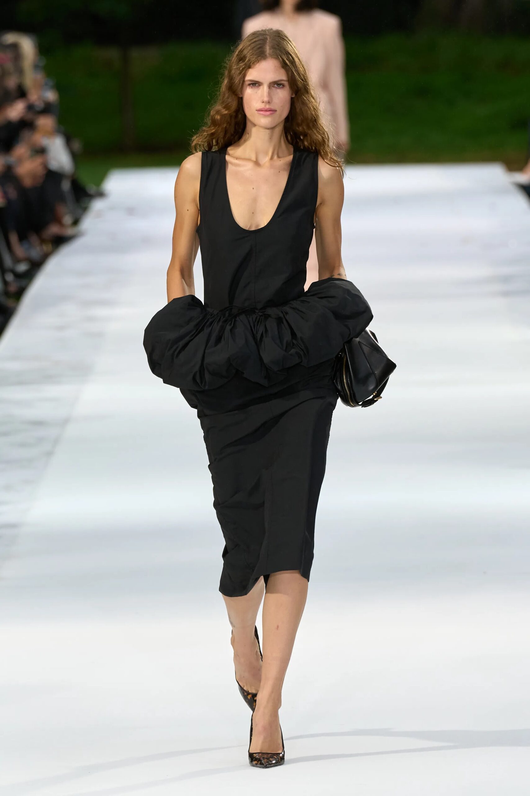 Modelo con un vestido negro de tirantes con detalles de volumen fruncido en la zona de la cintura y un bolso de mano oscuro.