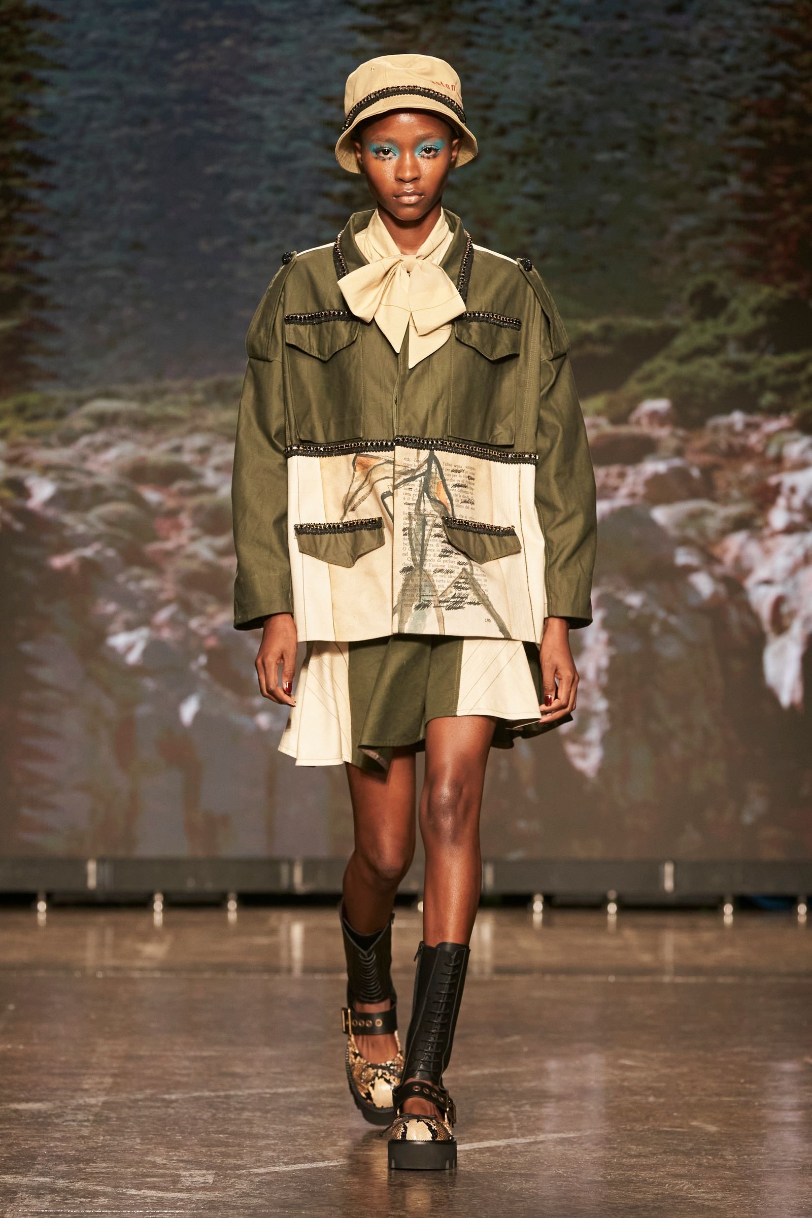 Modelo desfilando con un conjunto verde militar de chaqueta y falda con aplicaciones artísticas, sombrero tipo bucket y botas oscuras.