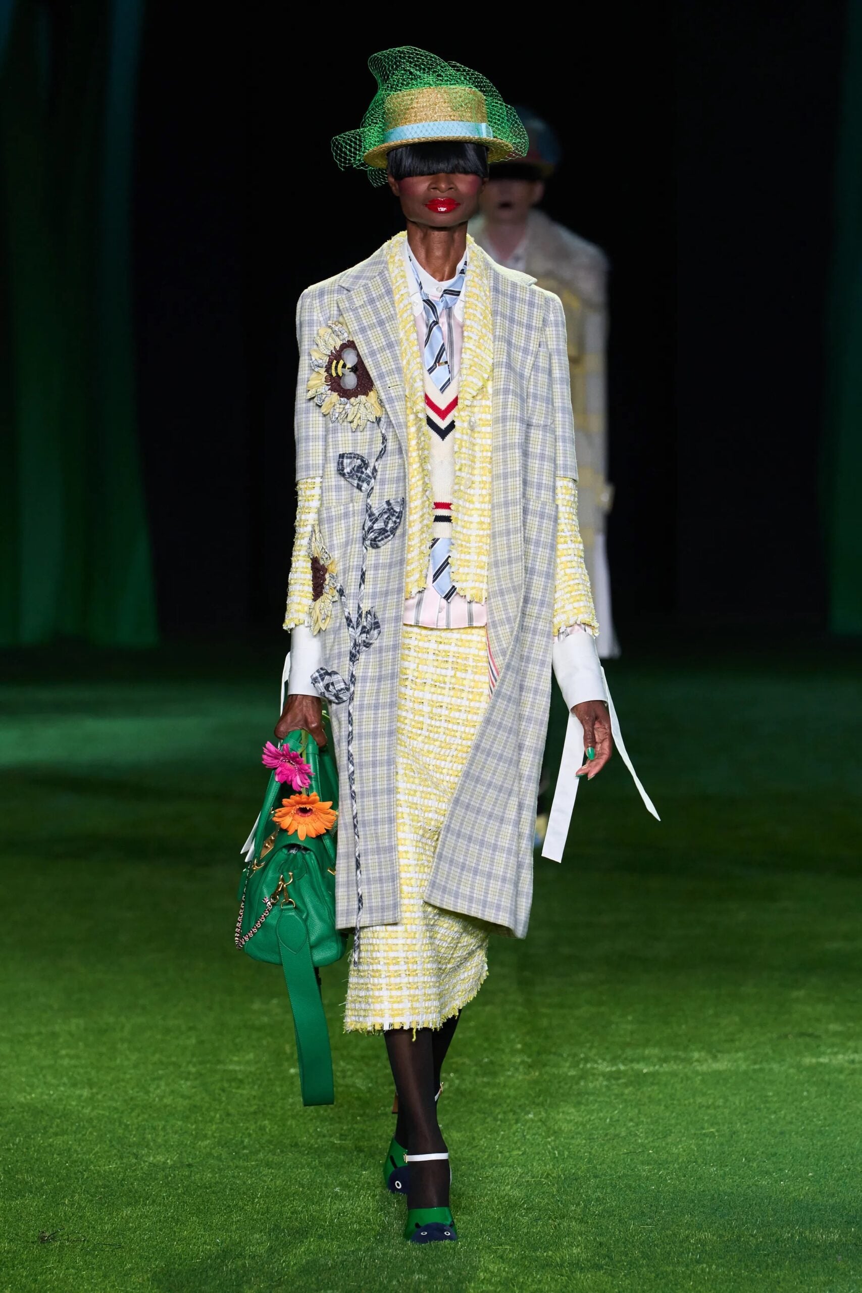 Modelo luciendo un abrigo largo de cuadros grises y amarillos con aplicaciones bordadas de girasoles, falda a juego y sombrero verde con red.