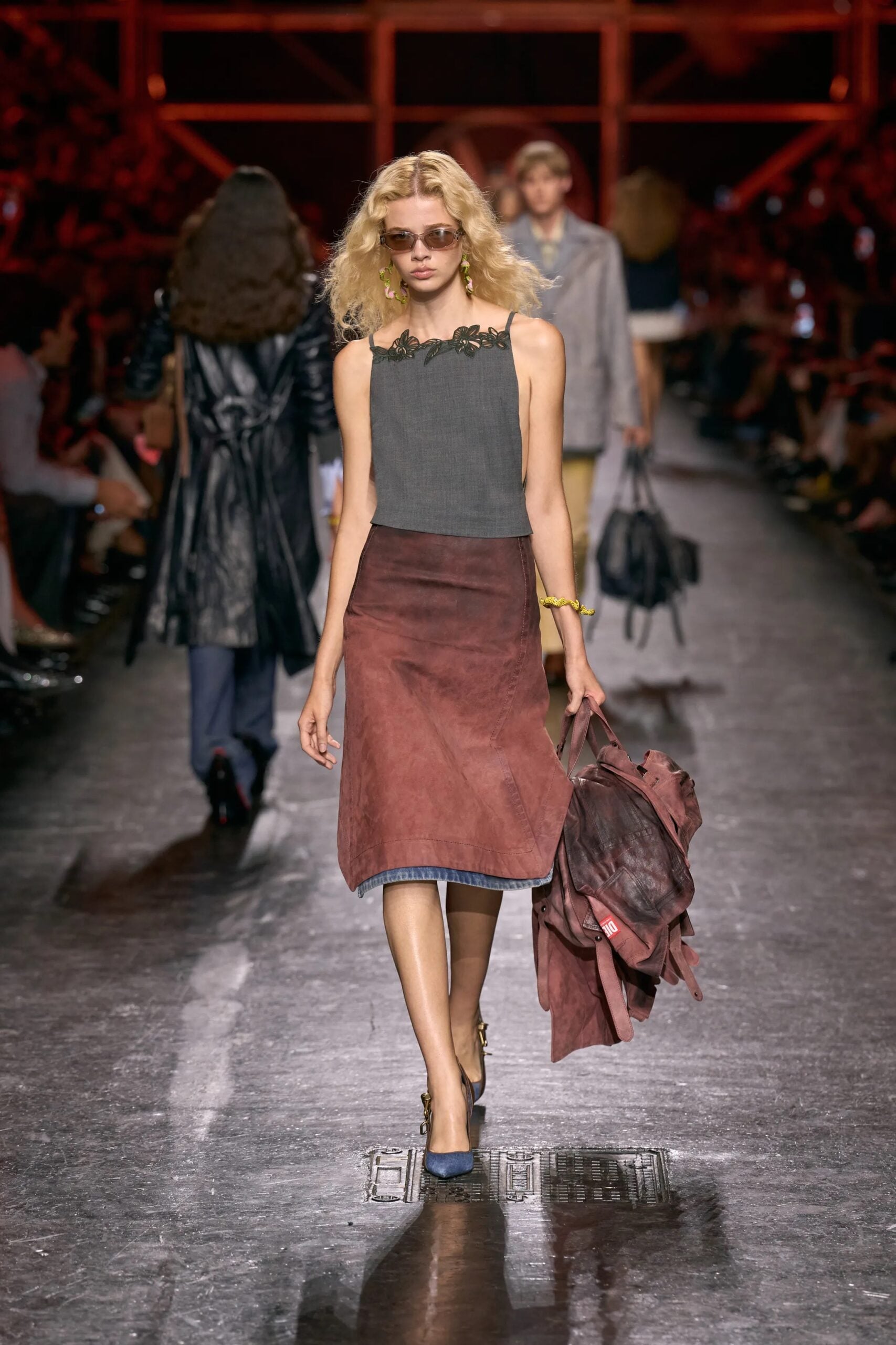 Modelo portando una blusa sin mangas gris con detalles decorativos en el escote, falda midi en tono rojizo con efecto deslavado y accesorios oscuros.