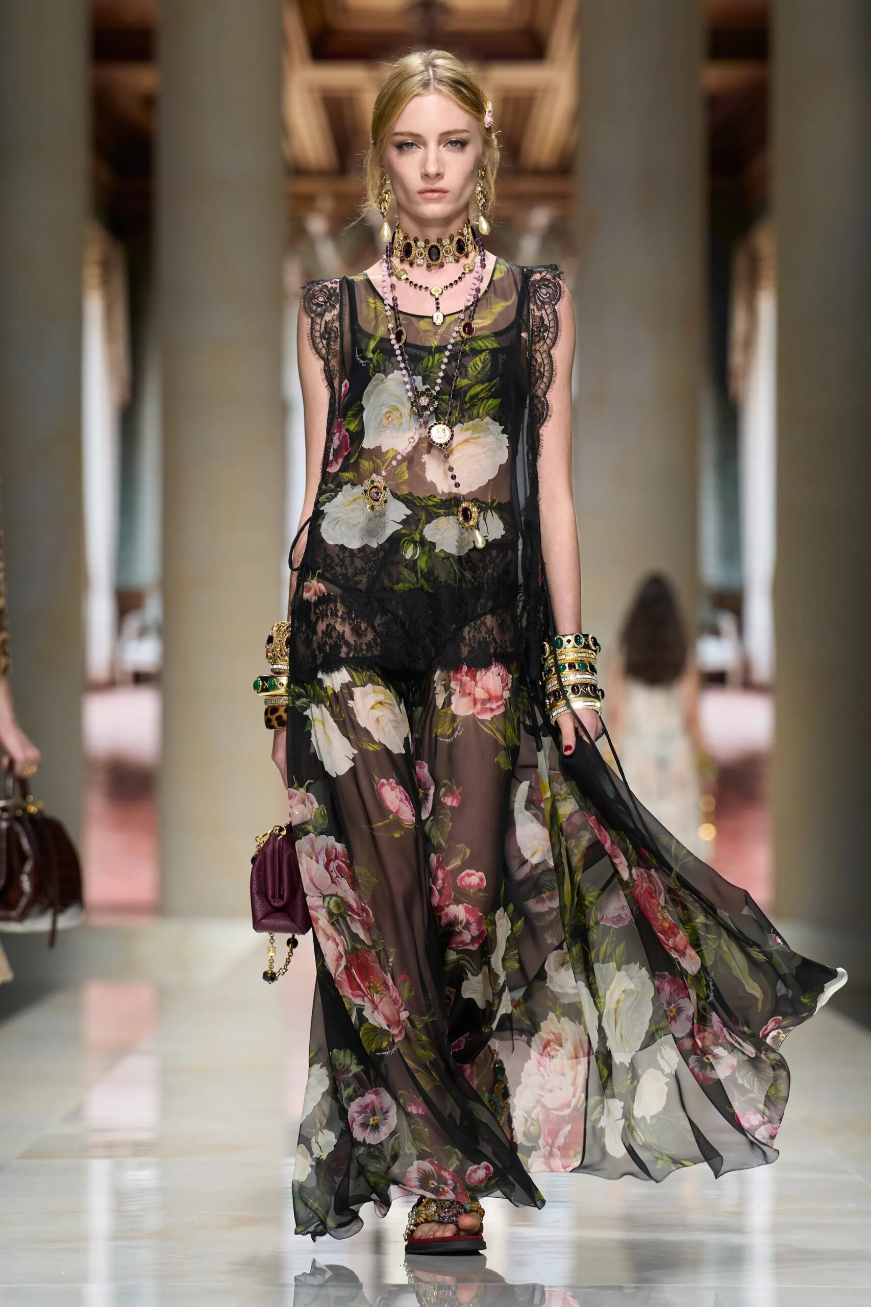 Modelo con un vestido largo y fluido de gasa negra con un vibrante estampado floral de rosas rojas y blancas, acompañado de joyería dorada ornamentada, fotografiada por Isidore Mantag