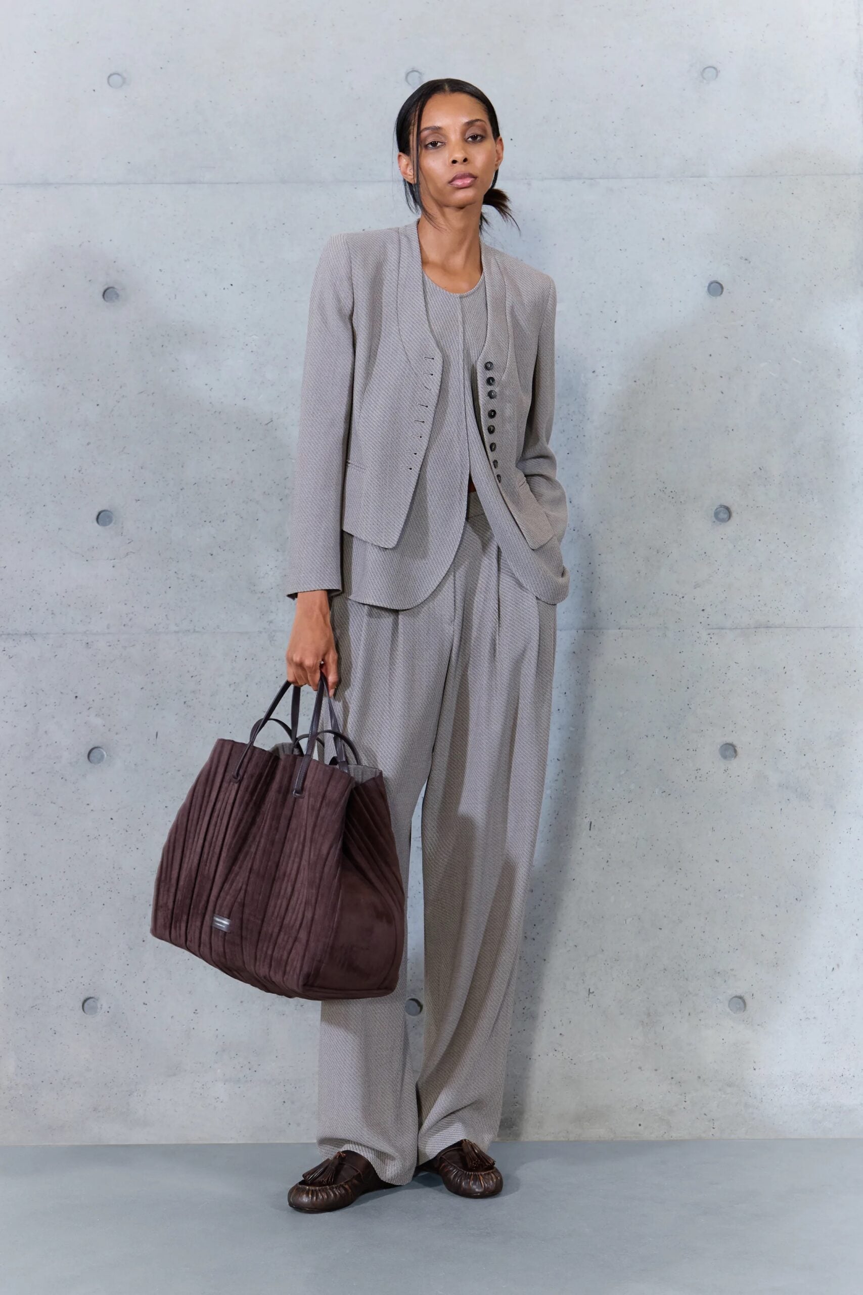 Modelo luciendo un conjunto sastre gris estructurado de chaqueta entallada y pantalón fluido, complementado con un bolso tote grande de ante marrón oscuro y zapatos planos.
