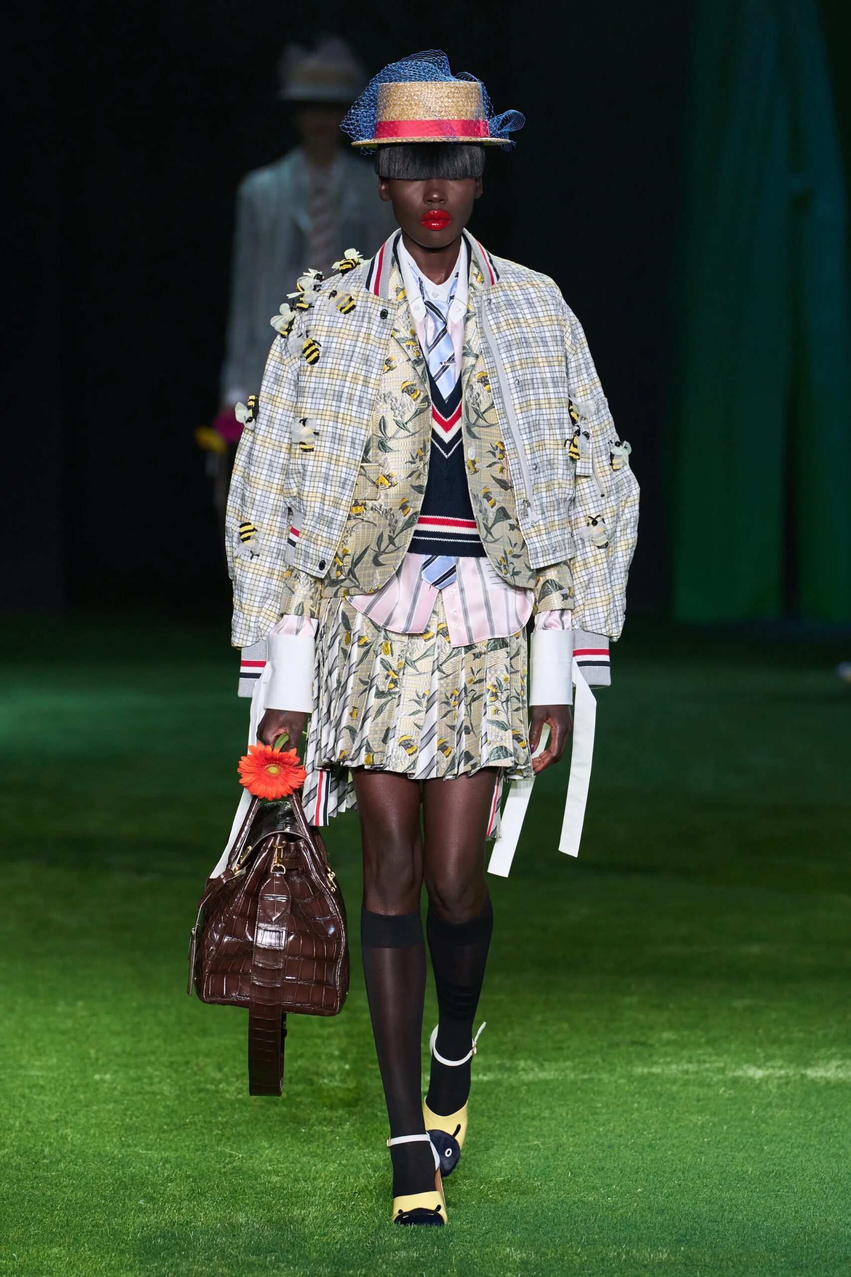 Modelo con una chamarra bomber estampada con motivos de abejas, falda plisada a juego, corbata y sombrero canotier con red azul.
