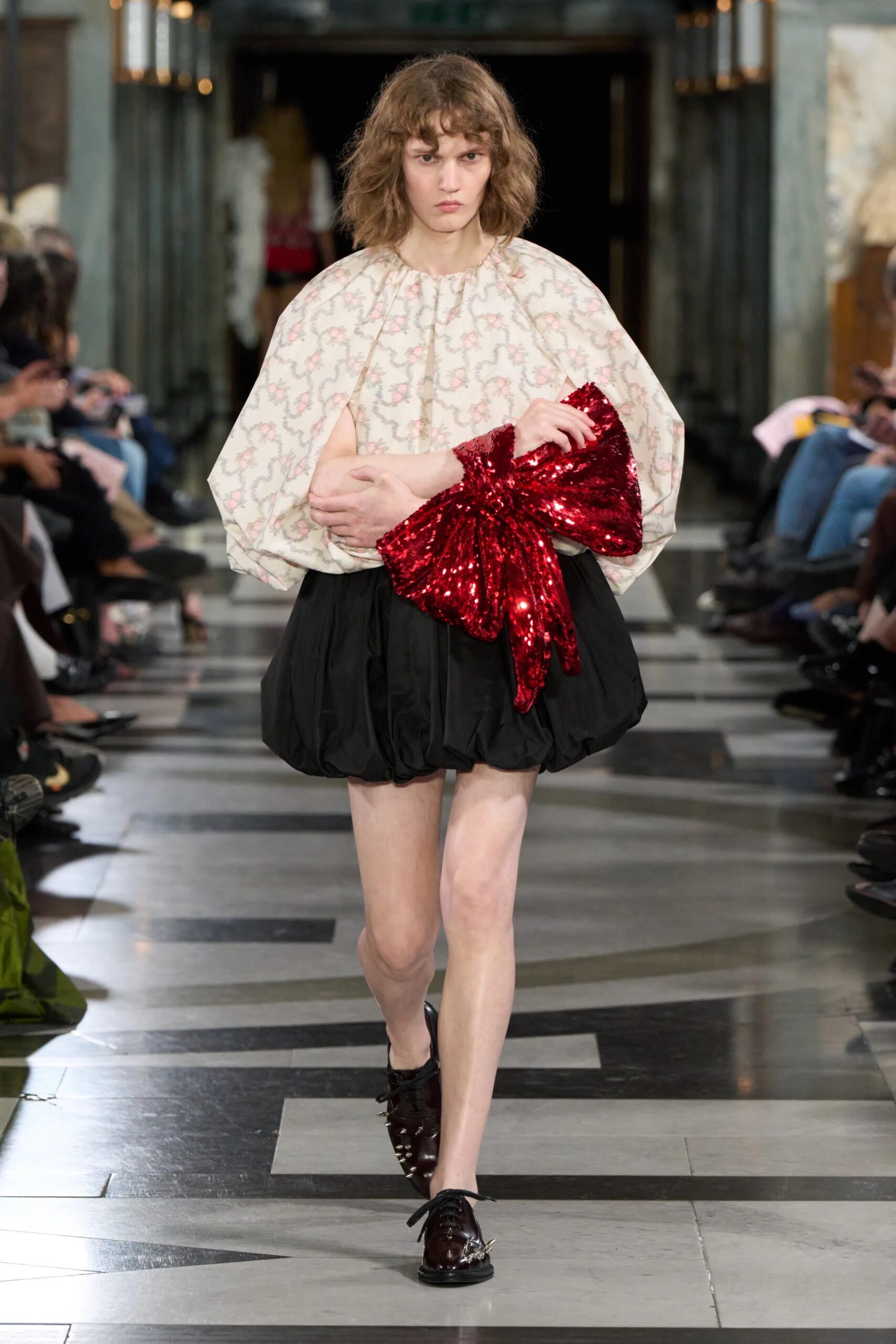 Modelo portando una blusa abombada con estampado floral suave, una falda corta negra con volumen y un llamativo lazo rojo de lentejuelas