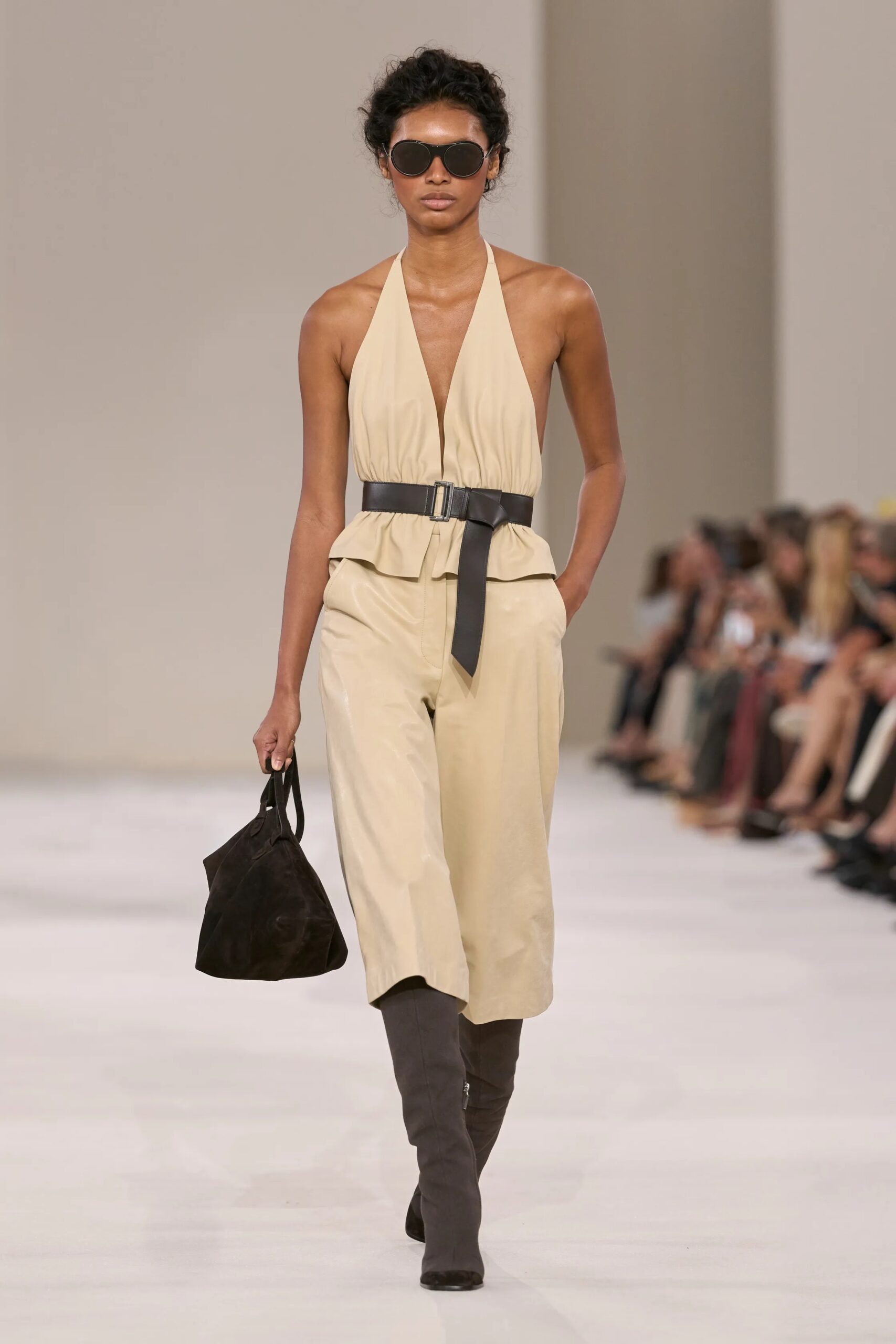 Modelo luciendo un conjunto de escote halter en tono beige con cinturón oscuro contrastante, bermudas a juego, gafas de sol y un bolso de mano negro.