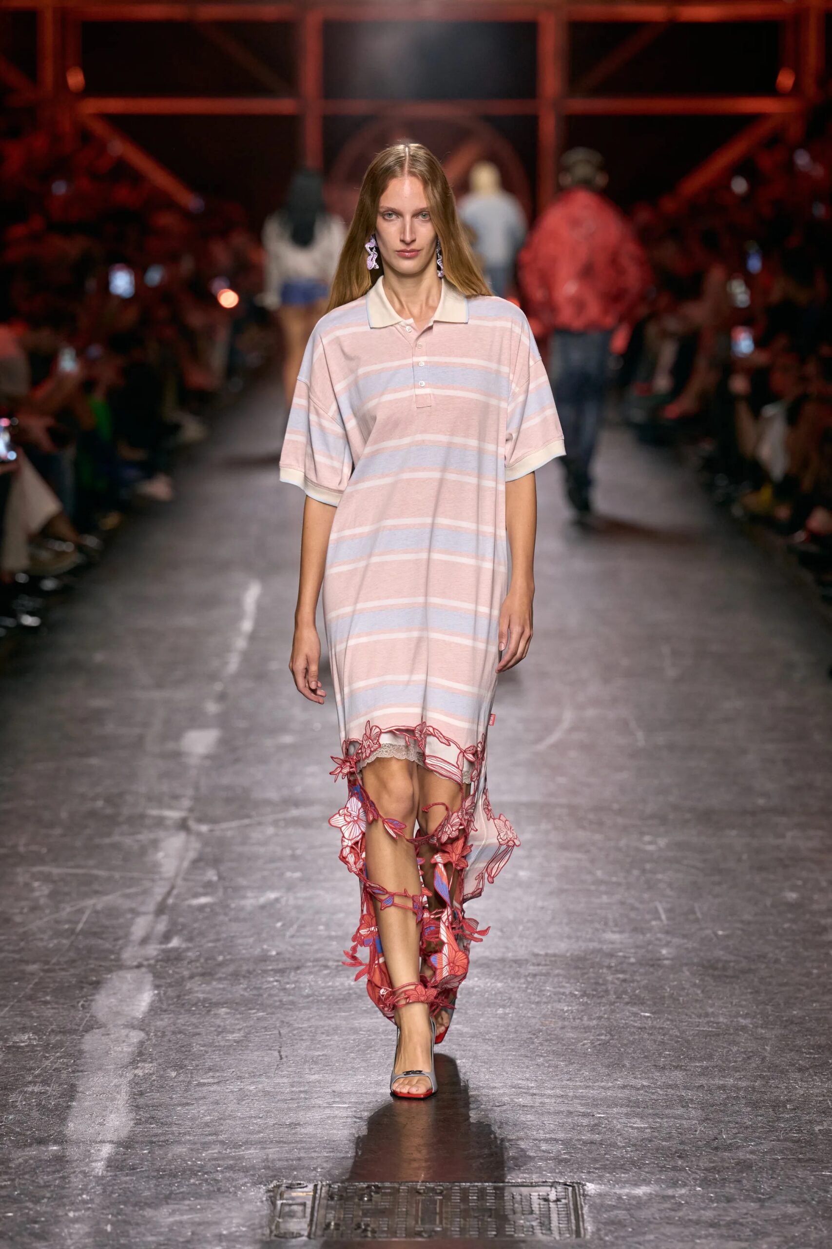 Modelo luciendo un vestido tipo polo con rayas horizontales en tonos rosados y celestes, con el bajo deshecho y adornado con aplicaciones colgantes.