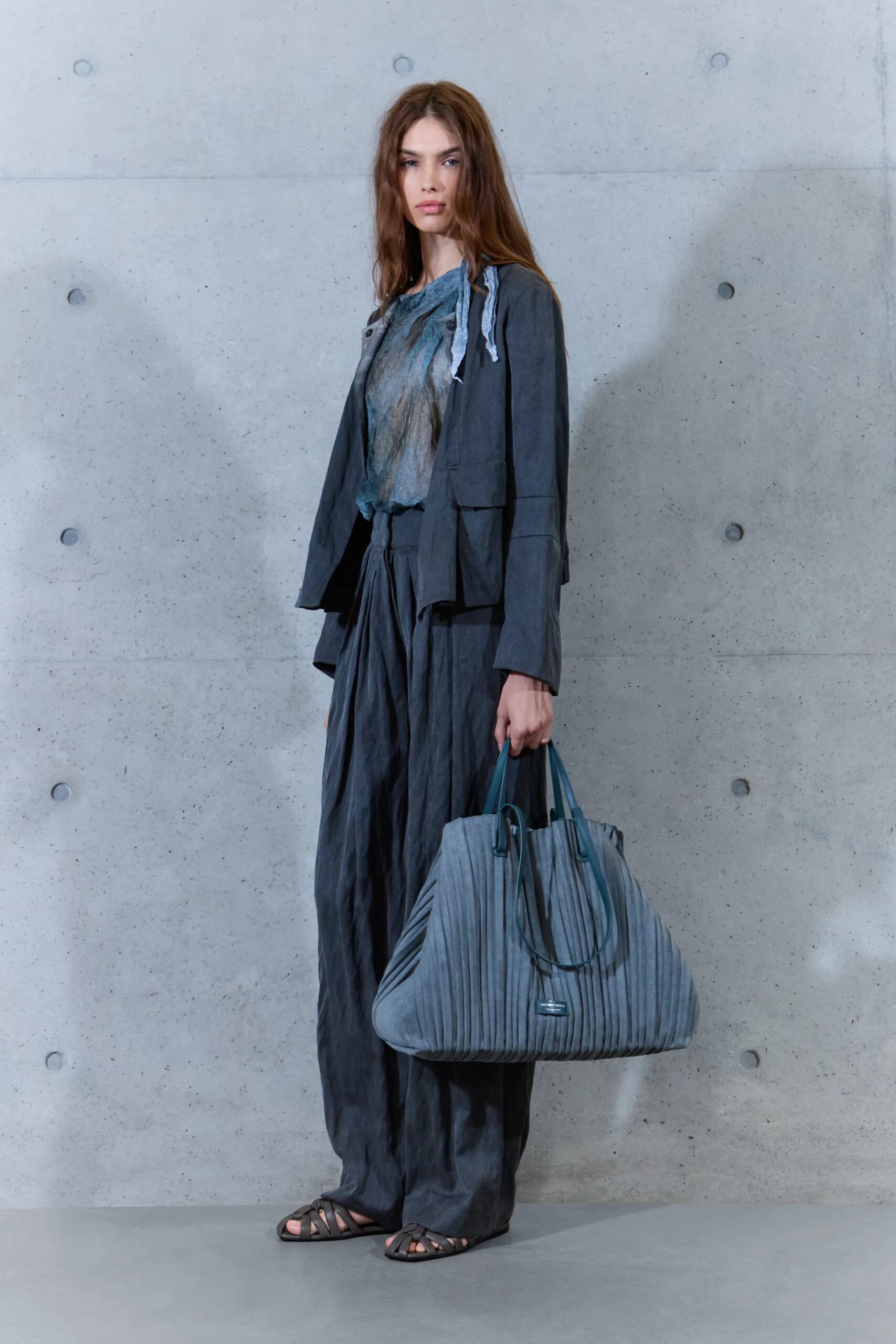 Modelo con un conjunto en tonos gris carbón de chaqueta ligera y pantalón amplio, portando un bolso tote plisado en el mismo tono y sandalias planas.