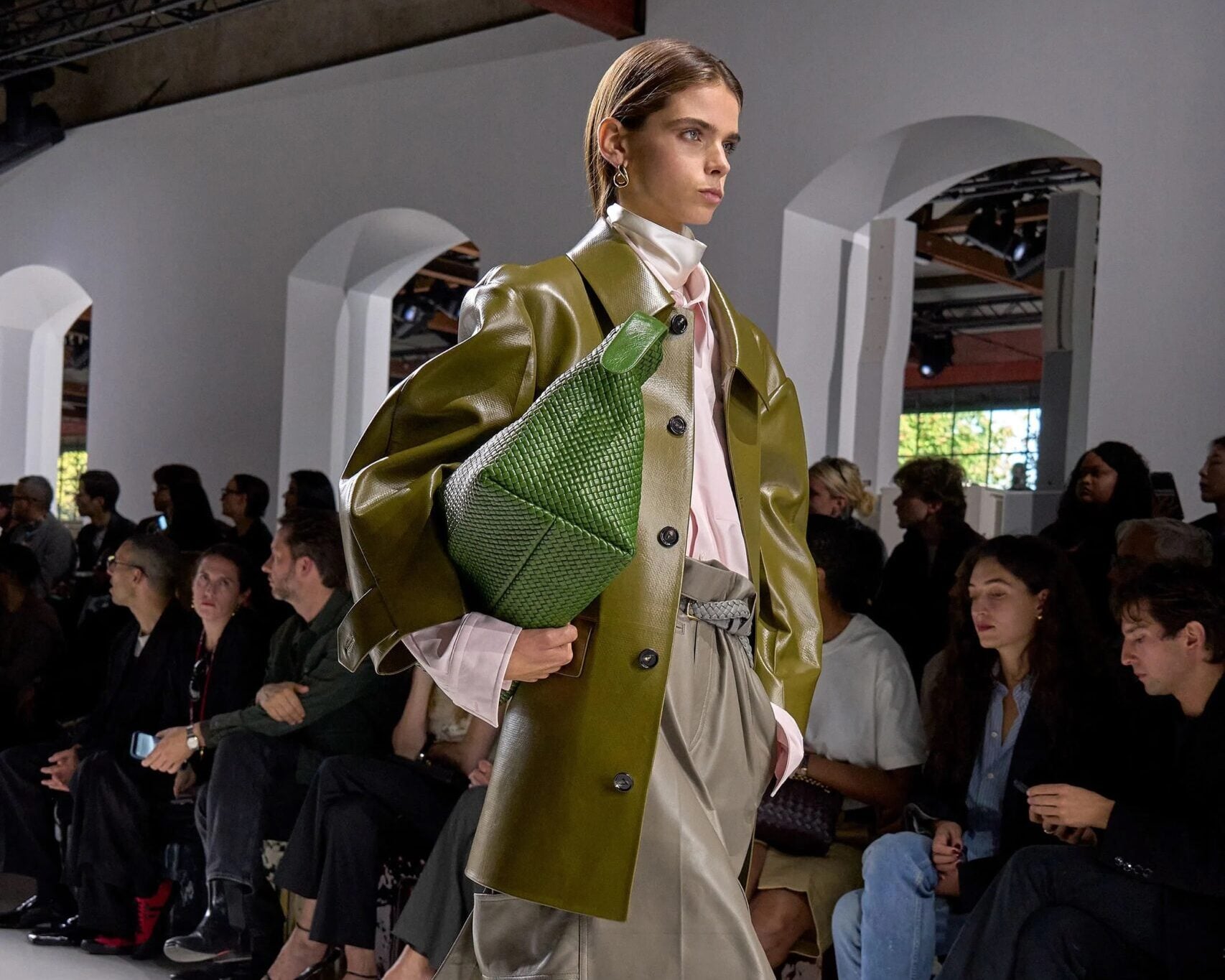 Modelo portando una chaqueta de piel en tono verde oliva, falda midi gris, un bolso tejido grande y zuecos claros en la pasarela.