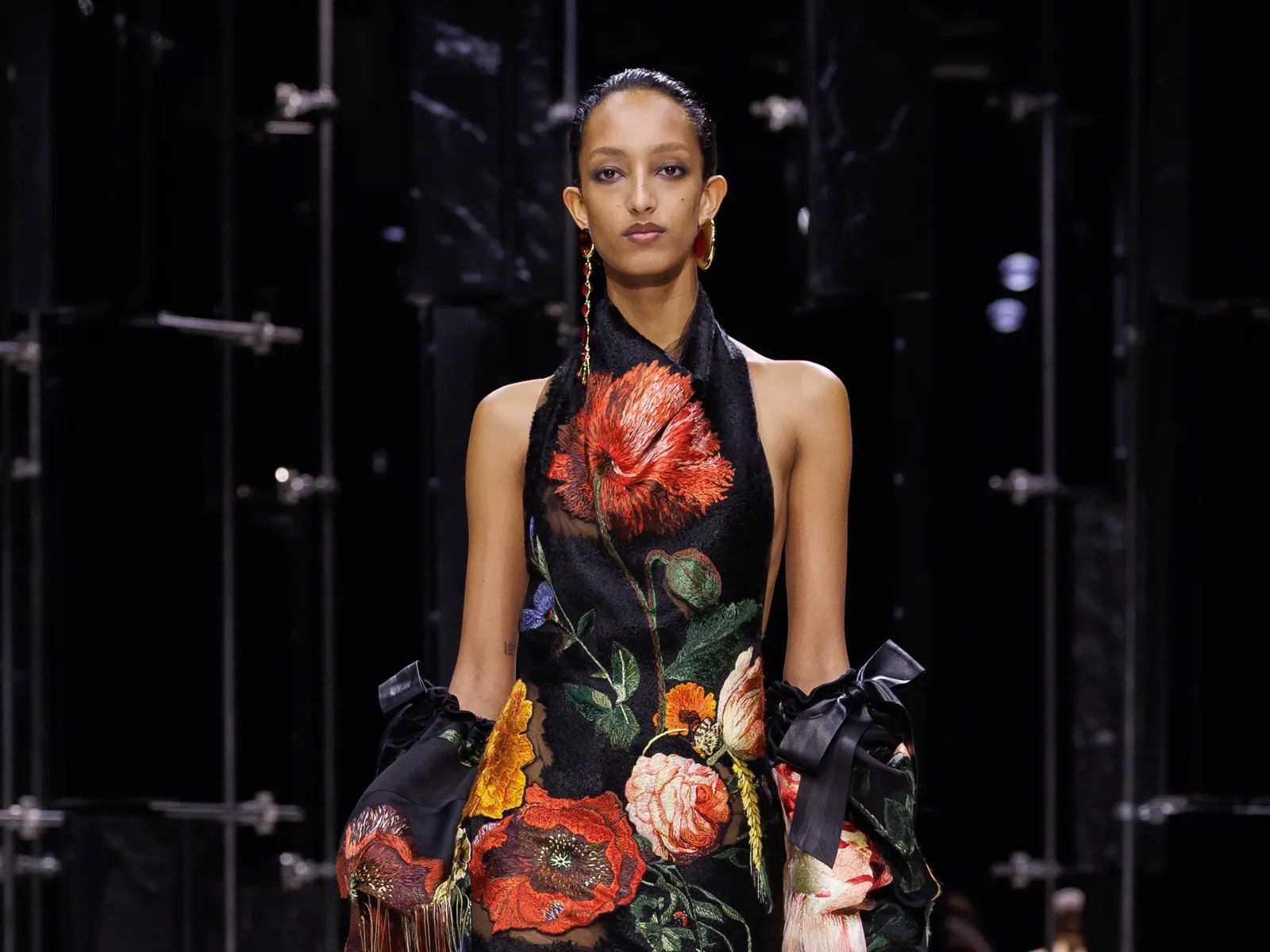 Primer plano de una modelo con un vestido negro halter adornado con grandes bordados florales bordados en tonos rojos y naranjas, y mangas caídas con lazos.