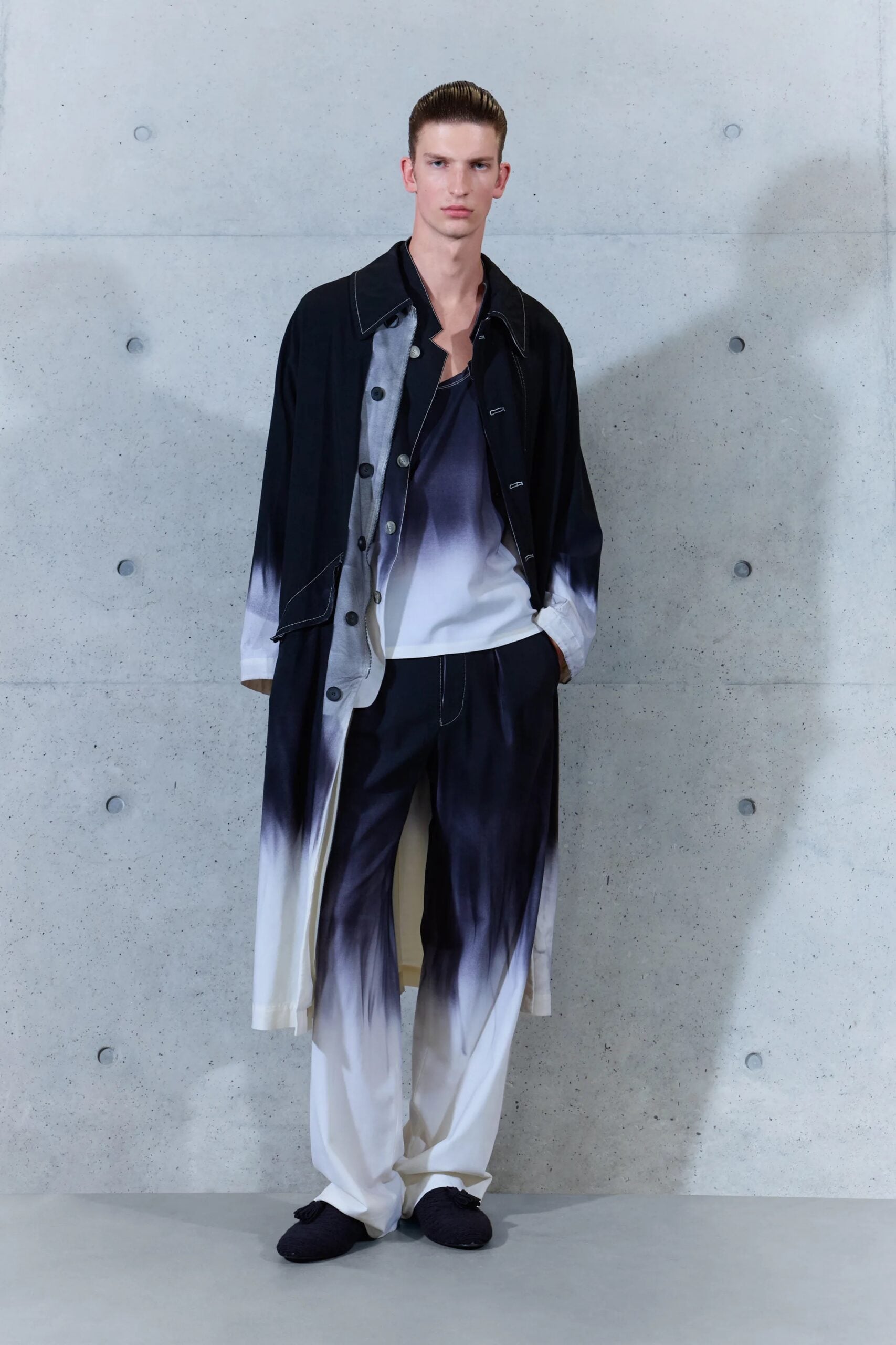 Modelo masculino portando un abrigo largo oscuro con efecto deslavado en degradé blanco, camisa a juego y pantalones amplios oscuros.