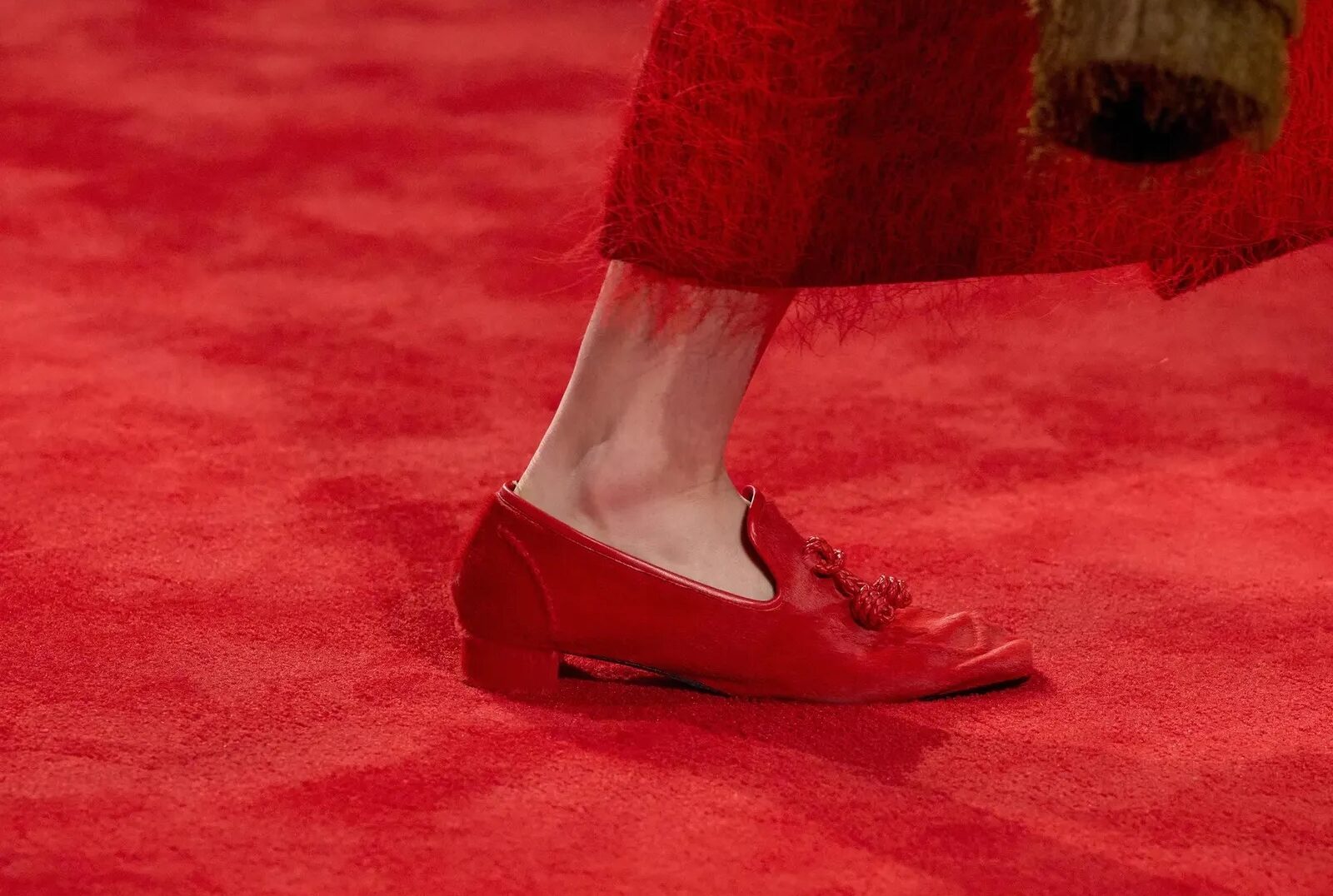 Detalle en pasarela de un mocasín rojo de piel con borlas coordinadas, combinado con una falda texturizada de flecos rojos sobre alfombra a juego.