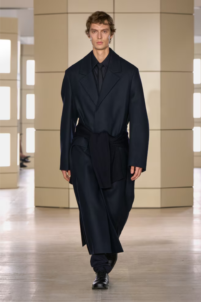 Modelo masculino con un abrigo largo cruzado en tono azul marino oscuro, camisa y corbata a juego, con un suéter amarrado a la cintura.