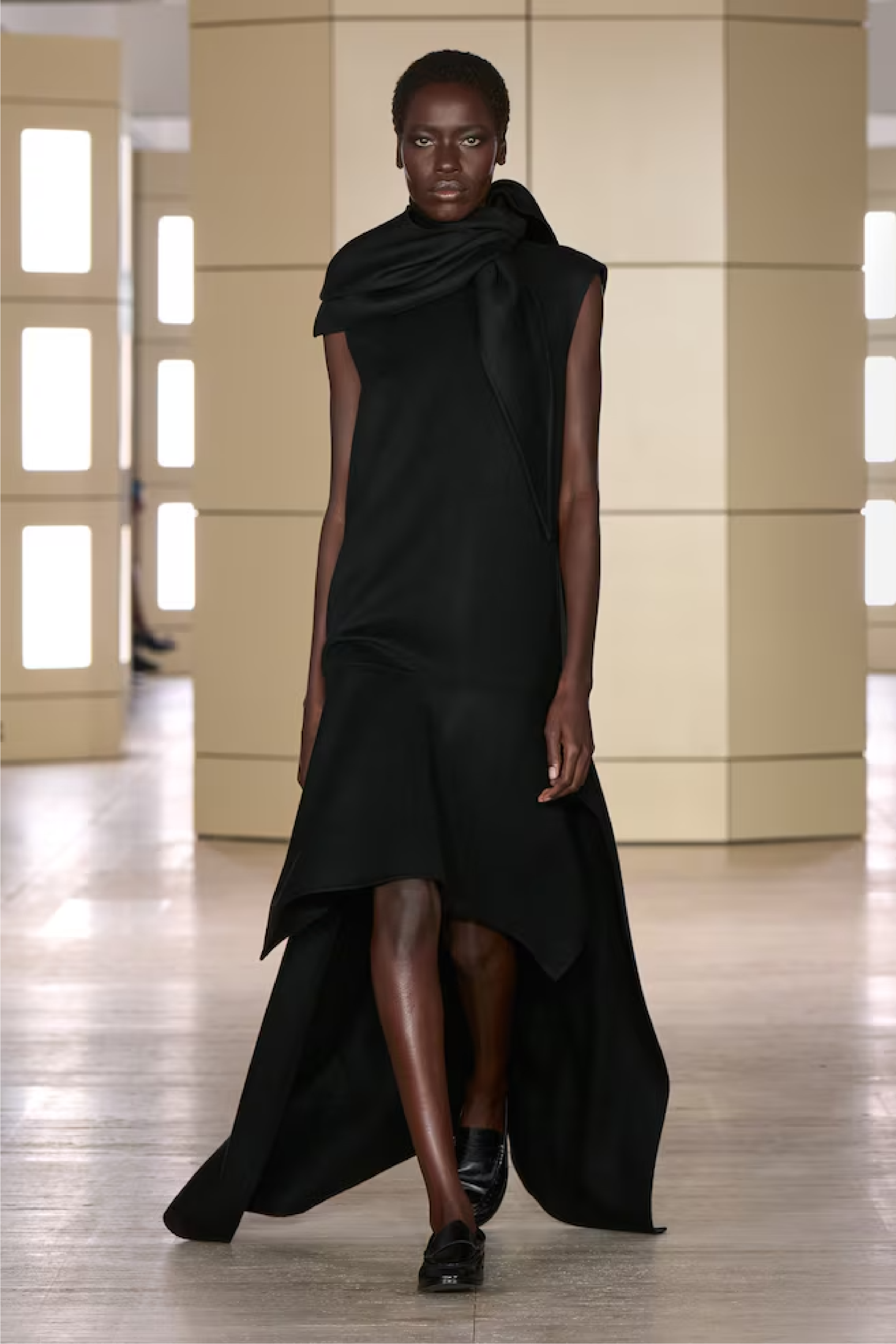 Modelo con un vestido largo negro sin mangas de corte asimétrico, bajo con vuelo y un foulard drapeado al cuello.
