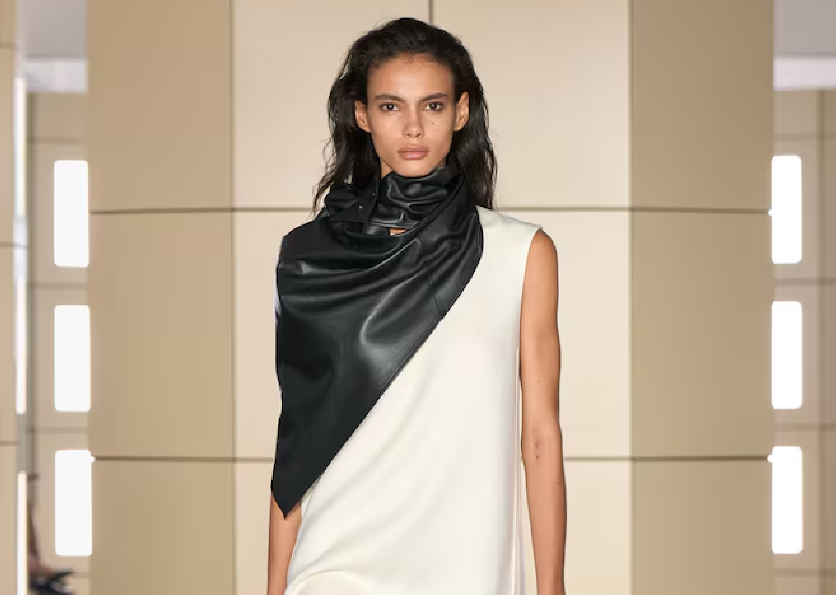 Modelo con un vestido blanco marfil sin mangas, bajo asimétrico y un estola o cuello de cuero negro drapeado sobre el hombro.