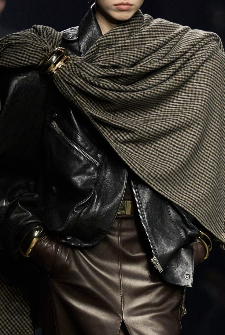 Primer plano de una chaqueta de piel negra combinada con una bufanda estampada de microcuadros drapeada sobre los hombros y sujeta con un broche metálico dorado.