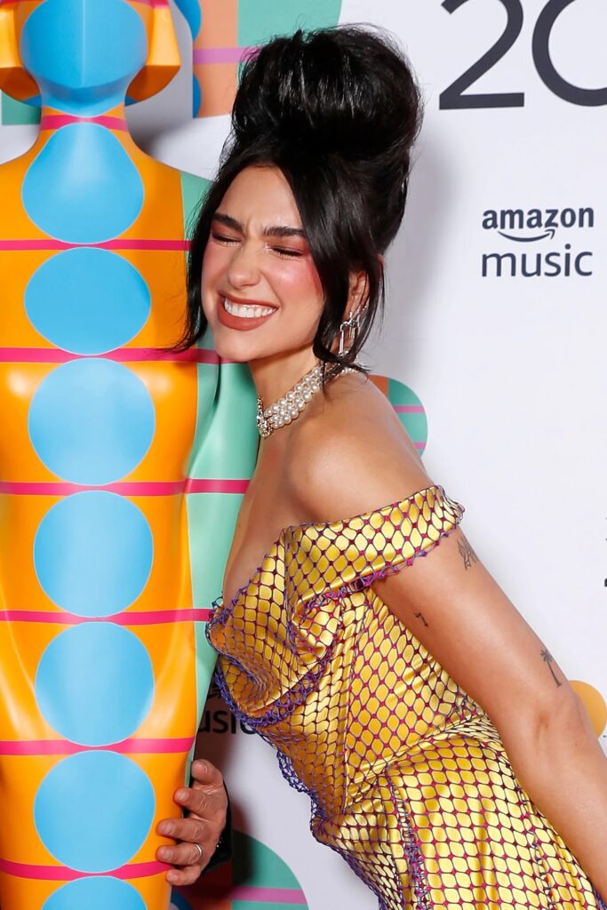 Dua Lipa sonriendo con un peinado voluminoso tipo beehive recogido en la coronilla, mechones sueltos al frente y un vestido dorado con detalles oscuros.
