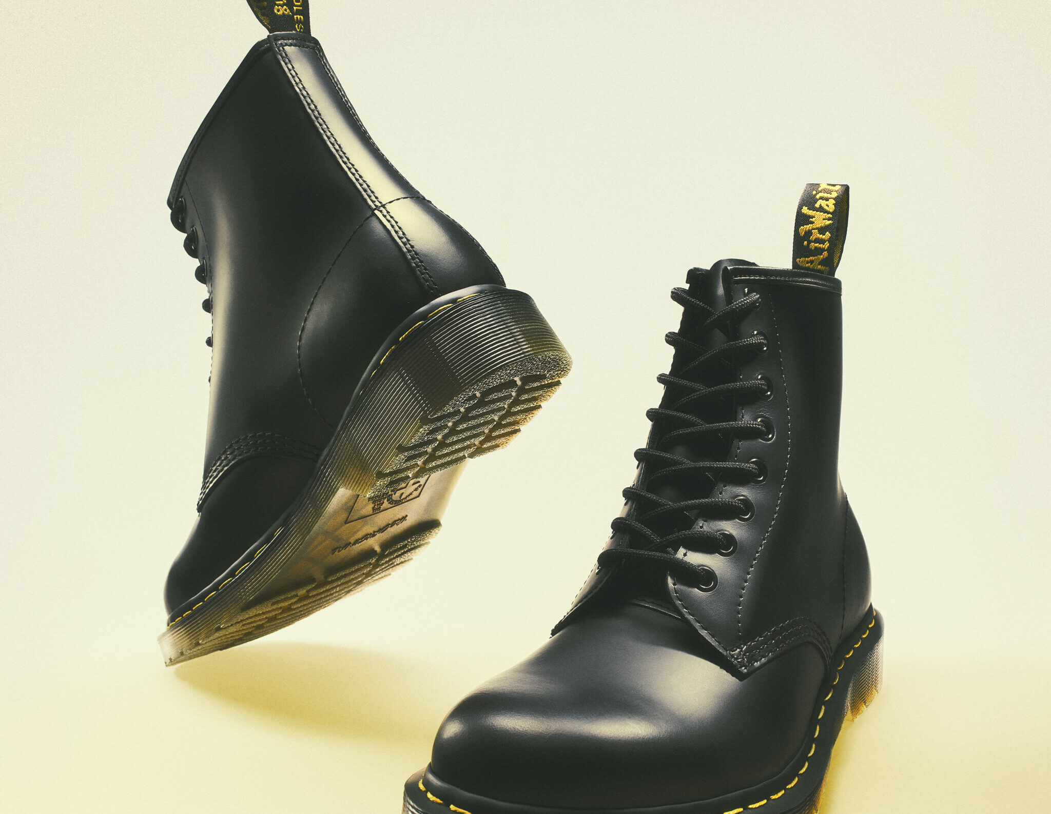 Par de botas Dr. Martens de piel negra dispuestas dinámicamente con una vista de suela y perfil sobre un fondo claro de estudio.