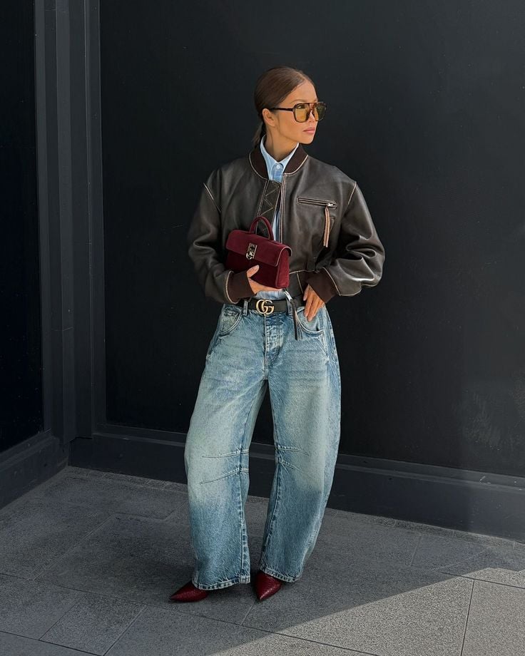 Mujer en la calle con una chaqueta bomber de cuero marrón, camisa azul, gafas de sol con cristales amarillos, un bolso de mano burdeos y jeans balloon claros.