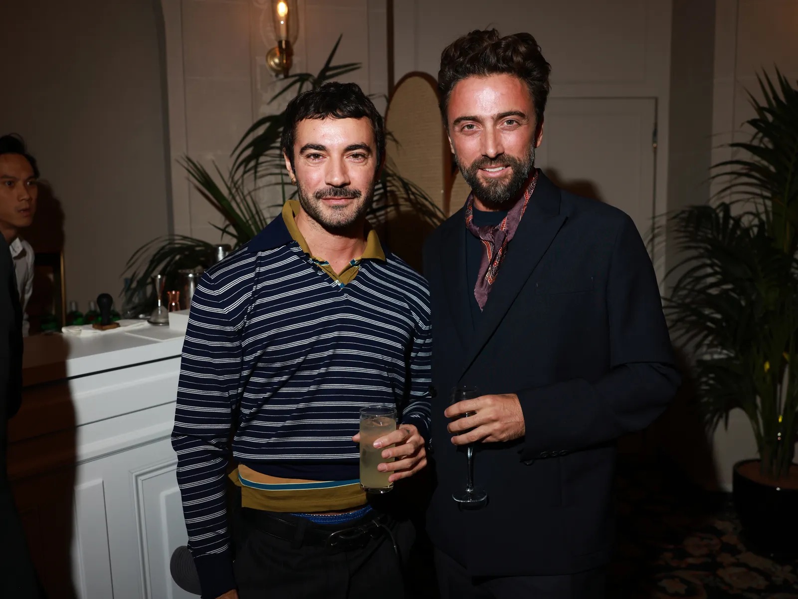 Dario Vitale de pie junto a otra persona en un evento social de interiores, vistiendo un suéter de rayas oscuras con cuello polo y sosteniendo un vaso con bebida.