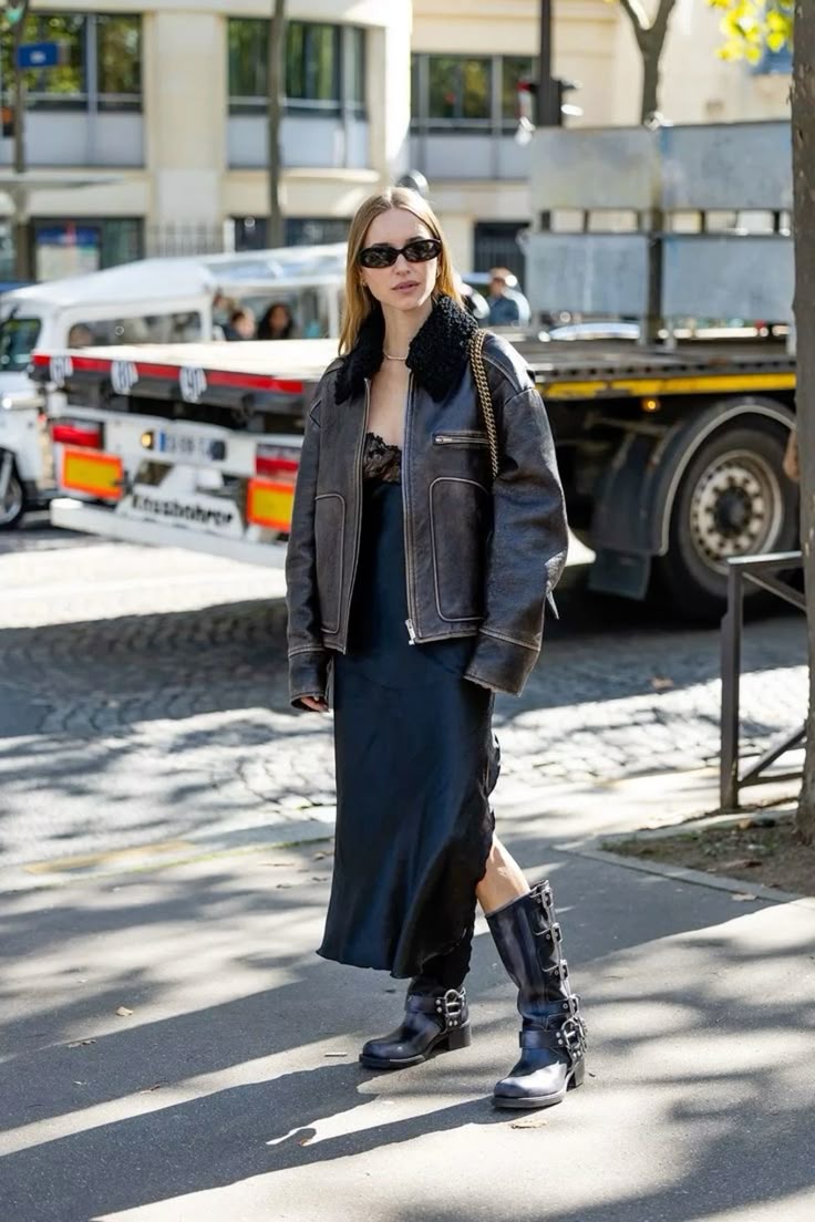Mujer caminando por la calle con una chaqueta de cuero desgastado con cuello de borrego, vestido midi negro satinado y botas biker negras altas con hebillas.