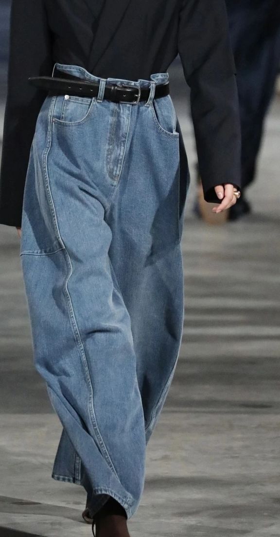 Vista de cerca de una modelo en pasarela con unos jeans balloon de talle alto ajustados con un cinturón negro y una chaqueta oscura estructurada.