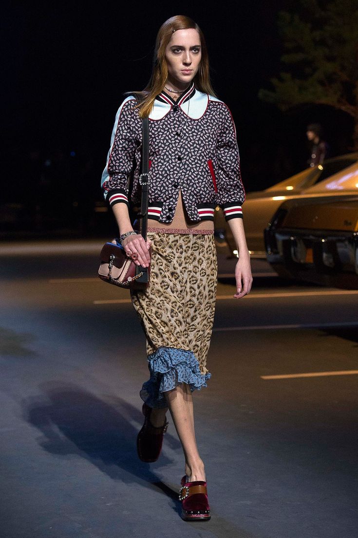 Modelo desfilando de noche en una pasarela al aire libre, vistiendo una chaqueta estampada de botones con cuello redondo, una falda midi con estampado de leopardo y detalles de volantes azules en el bajo, un bolso cruzado y zapatos oscuros.