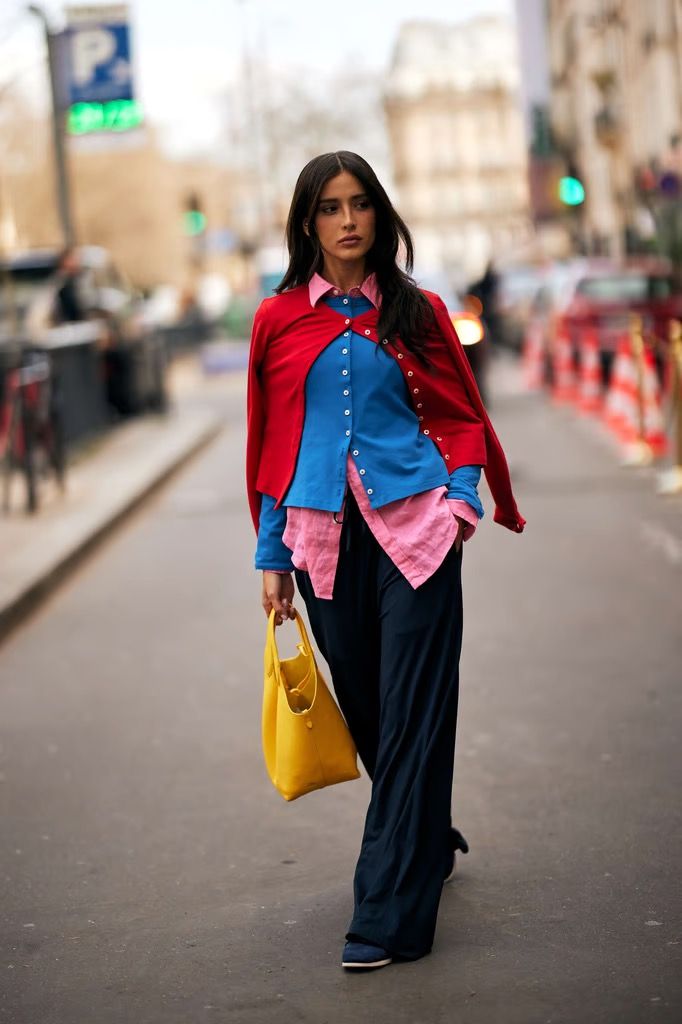 Mujer caminando por la calle luciendo un llamativo juego de capas con camisa rosa, camisa azul abotonada encima, cárdigan rojo sobre los hombros, pantalones fluidos oscuros y un bolso amarillo.