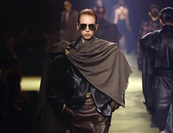 Modelo caminando por una pasarela con iluminación tenue, luciendo una chaqueta de cuero negro, una falda midi de piel marrón y una bufanda a cuadros dispuesta como echarpe con un broche dorado.