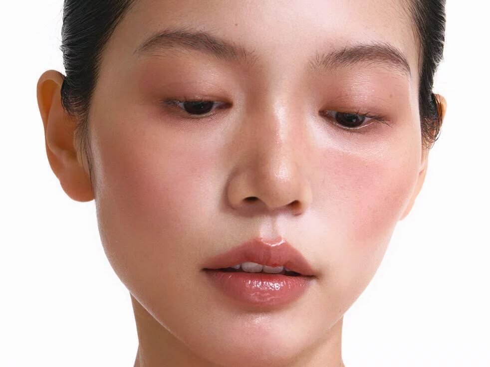Primer plano lateral de un rostro con piel de acabado ultra luminoso (efecto "glass skin"), mejillas con un rubor rosado difuminado y labios con brillo natural en tonos nude.