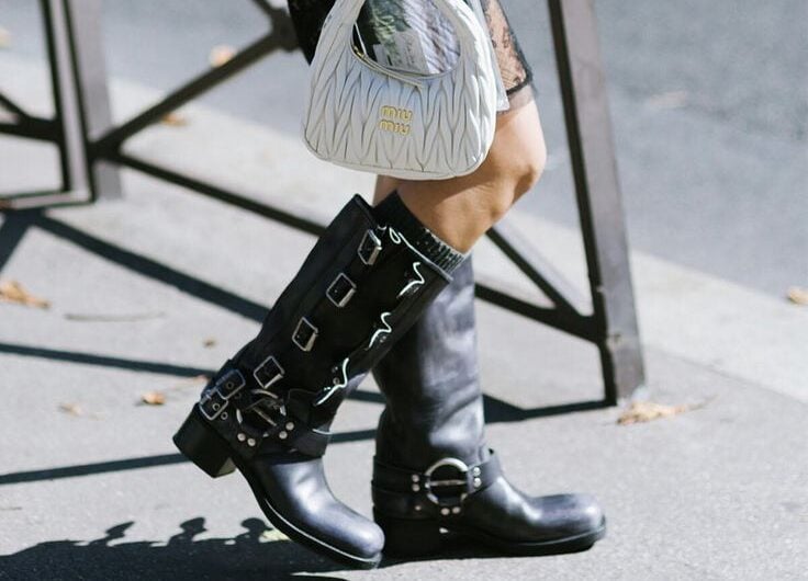 Detalle de las piernas de una mujer luciendo botas biker negras con múltiples hebillas, calcetines oscuros y un bolso pequeño blanco capitoné de mano.