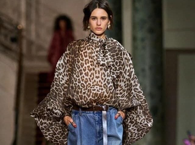 Modelo caminando sobre una pasarela con una blusa vaporosa de estampado de leopardo con lazo al cuello, jeans balloon clásicos y accesorios dorados.