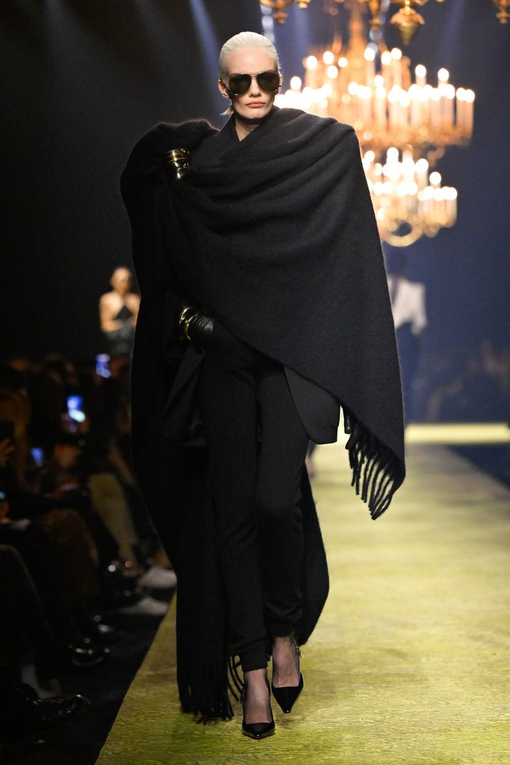Modelo con cabello rubio platino desfilando en la pasarela con un total look negro compuesto por pantalón, chaqueta oculta y una voluminosa bufanda negra drapeada como echarpe con flecos en los extremos.