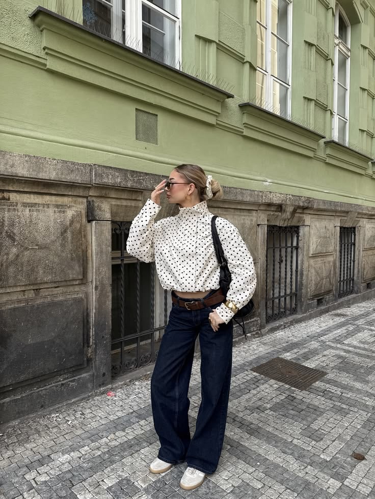 Mujer frente a un edificio verde con una blusa estructurada de cuello alto con estampado de lunares, jeans oscuros de corte recto y cinturón marrón.
