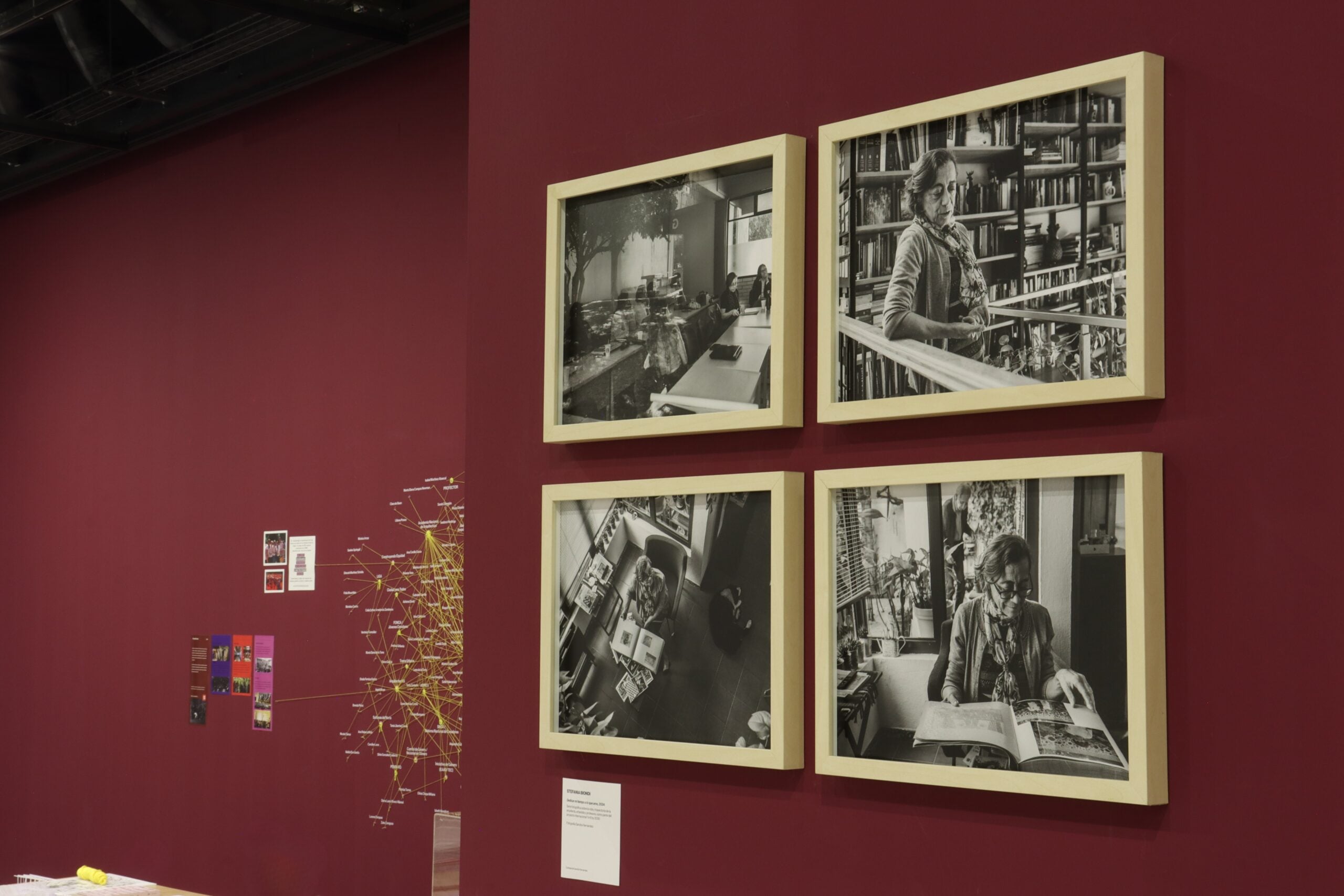 Muro de tono burdeos en una sala de exposición que exhibe cuatro fotografías enmarcadas en blanco y negro, mostrando espacios de trabajo y retratos de arquitectas, acompañado por un esquema conceptual tejido en la pared lateral.