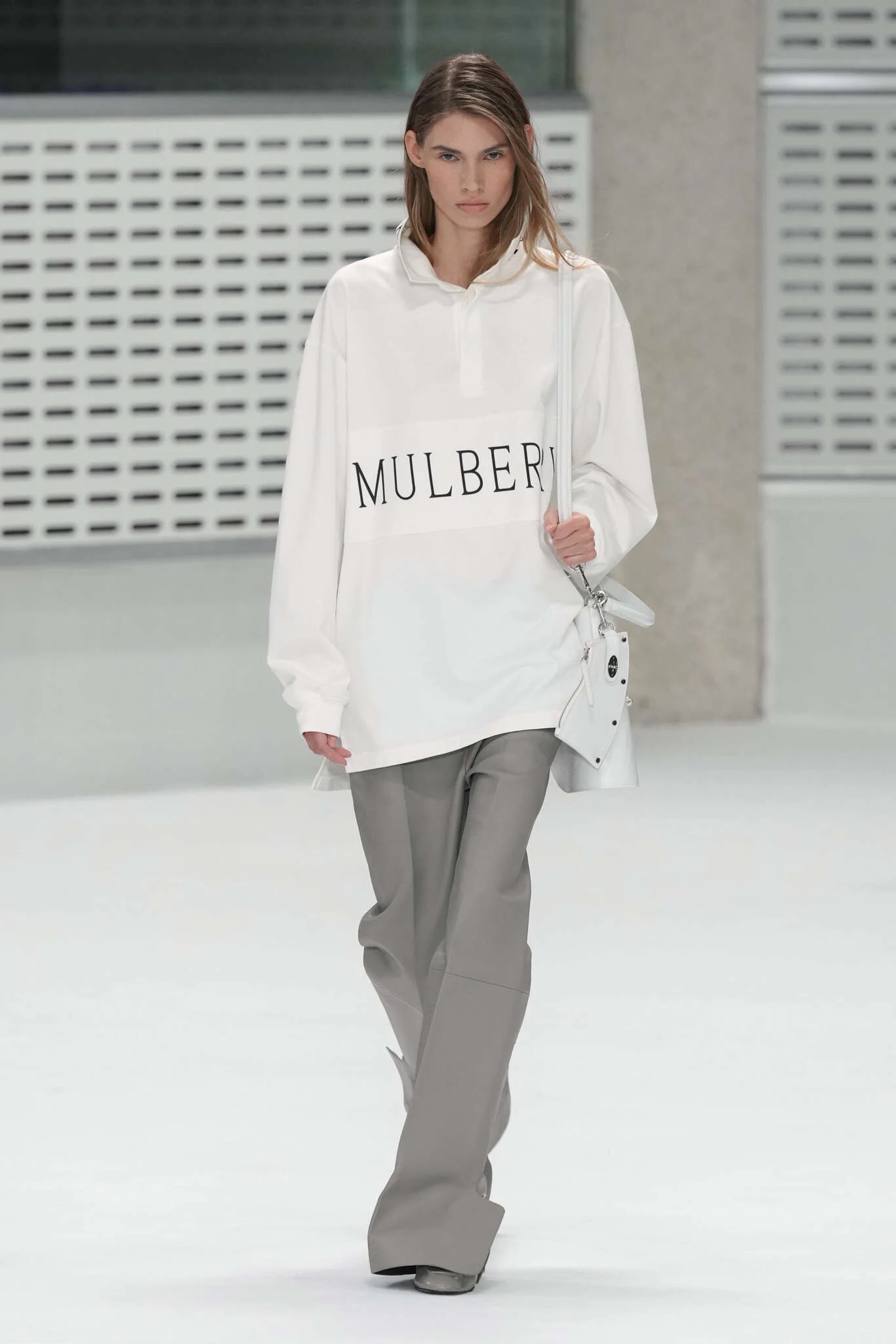 Modelo portando una sudadera deportiva blanca con el logotipo de Mulberry en contraste, pantalón fluido gris y un bolso cruzado.