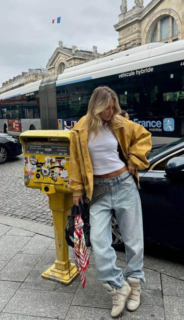 Mujer de pie en una calle urbana frente a un transporte público, luciendo una chaqueta de ante mostaza, camiseta blanca básica, jeans holgados y zapatillas con cuña oculta.