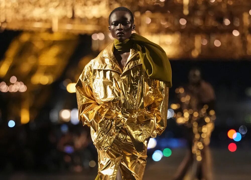 Modelo desfilando por la pasarela nocturna con un conjunto metalizado en tono dorado brillante de chaqueta estructurada y falda con abertura, acompañado de un fular verde oliva.