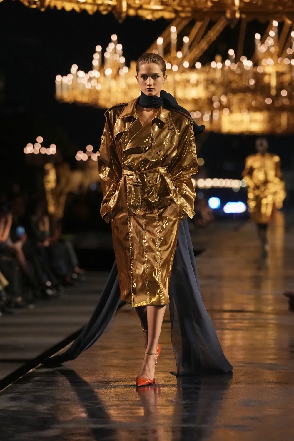 Modelo portando un abrigo trench metalizado en acabado dorado brillante con un cinturón ajustado, complementado con un fular oscuro y zapatos de tacón