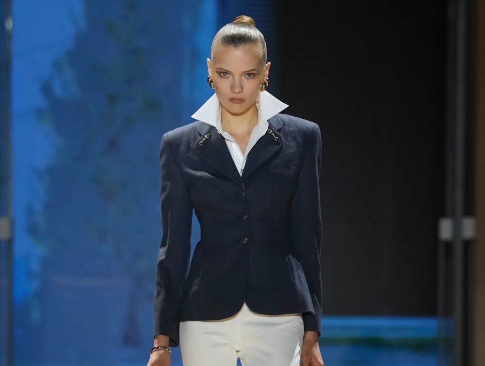 Modelo desfilando con una chaqueta estilo sastre en tono oscuro con cuello alto blanco estructurado, pantalones ajustados blancos y botas altas de piel negra, capturada por Alessandro Fucioni.