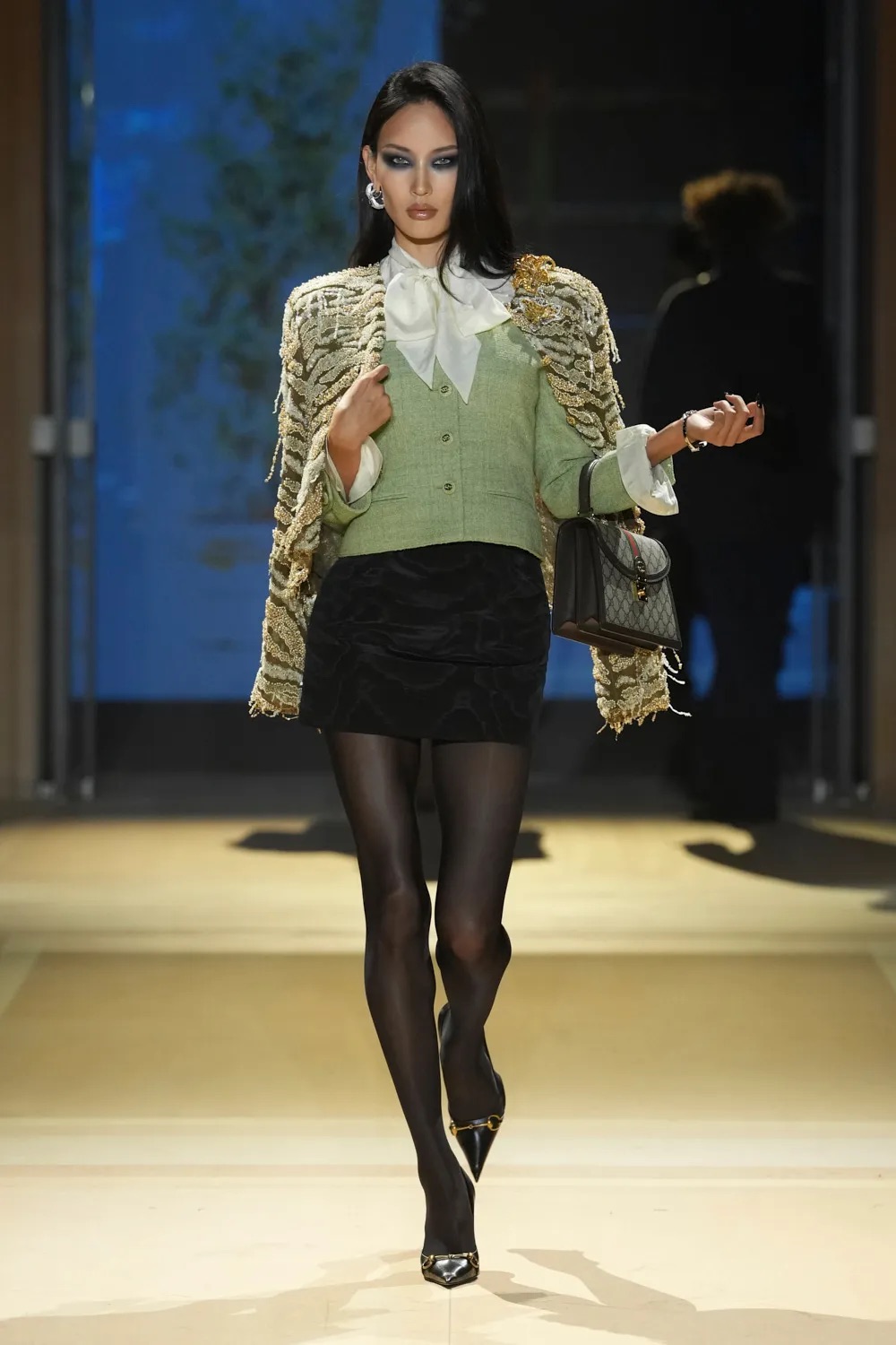 Modelo portando un cárdigan verde claro con detalles texturizados, una blusa blanca con lazo al cuello, falda corta negra, medias oscuras y zapatos de tacón, fotografiada por Alessandro Fucioni.