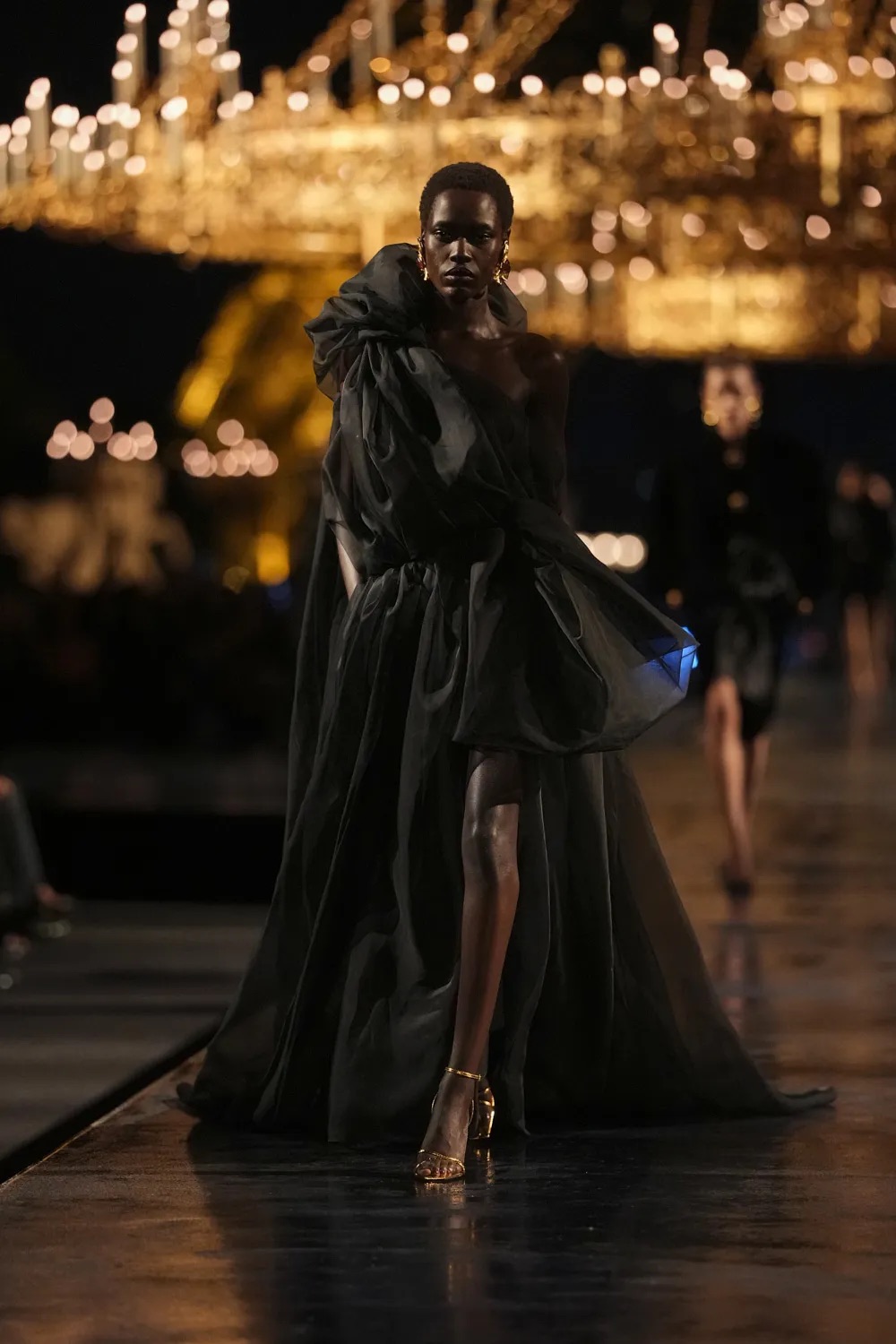 Modelo luciendo un vestido largo negro de alta costura con un cuerpo drapeado y voluminoso de organza transparente, con una abertura lateral que deja ver sus piernas.